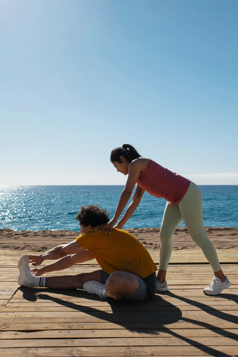 Woman assisting man stretching his legs on a wooden deck by the ocean on a sunny day.