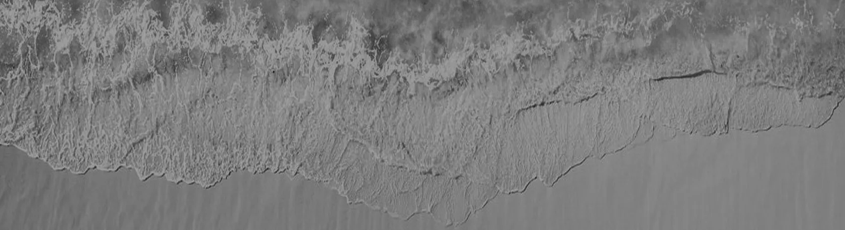 Black and white aerial view of ocean waves gently breaking onto a sandy shore.