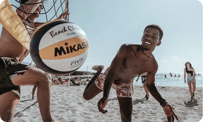 Two men playing beach volleyball near the net on a sunny sandy beach with people in background.