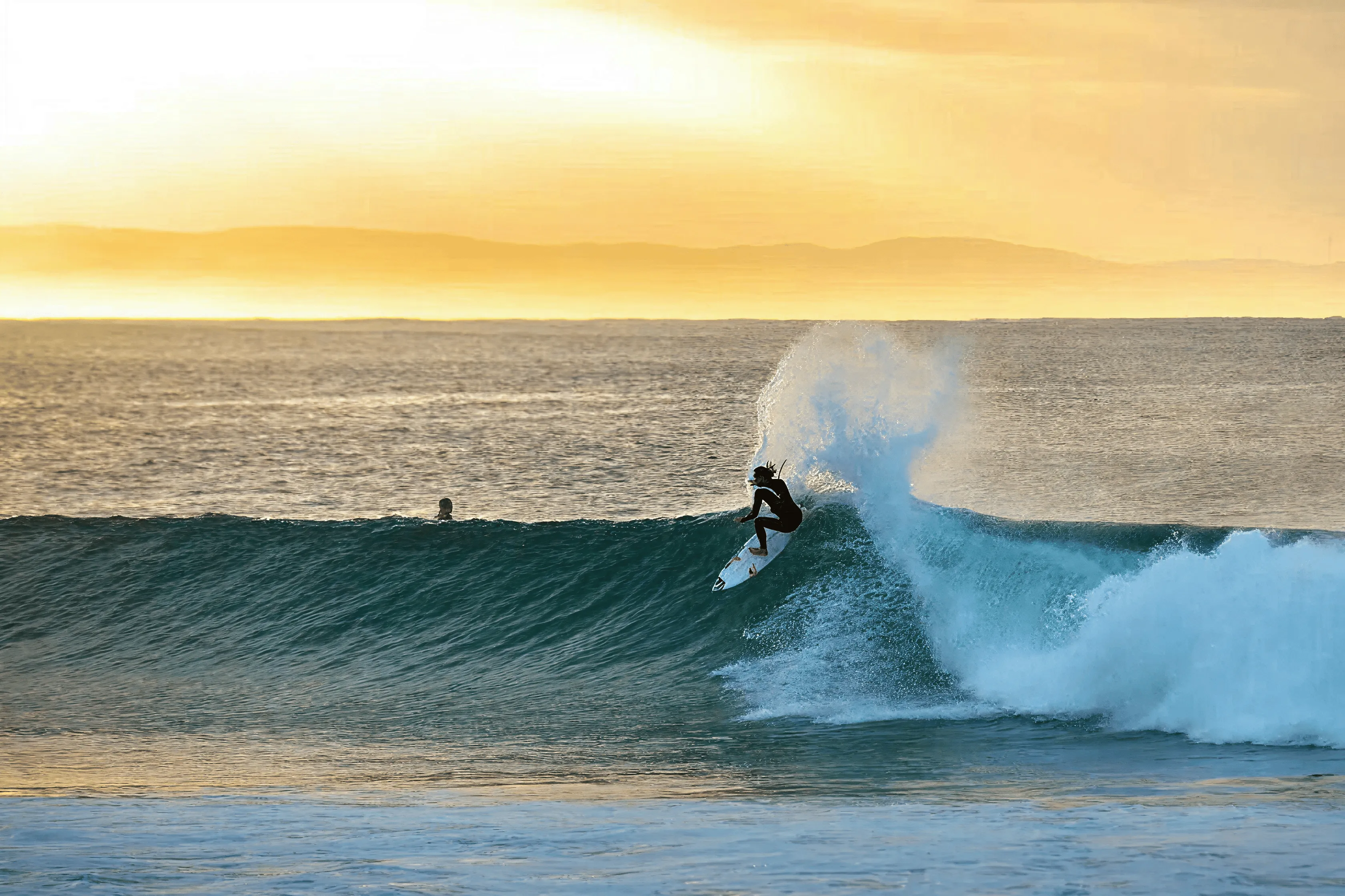 Surfer riding a wave with spray under a golden sunset over the ocean and distant hills.