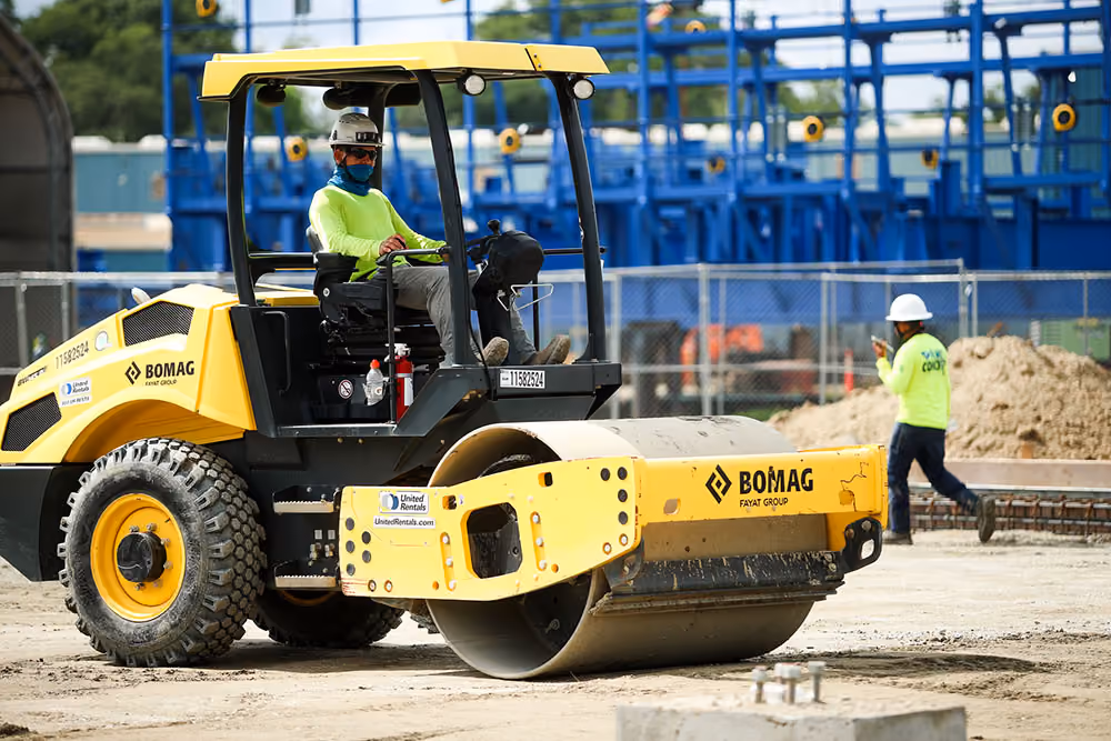 Construction worker operating yellow BOMAG roller on dirt surface at active construction site.