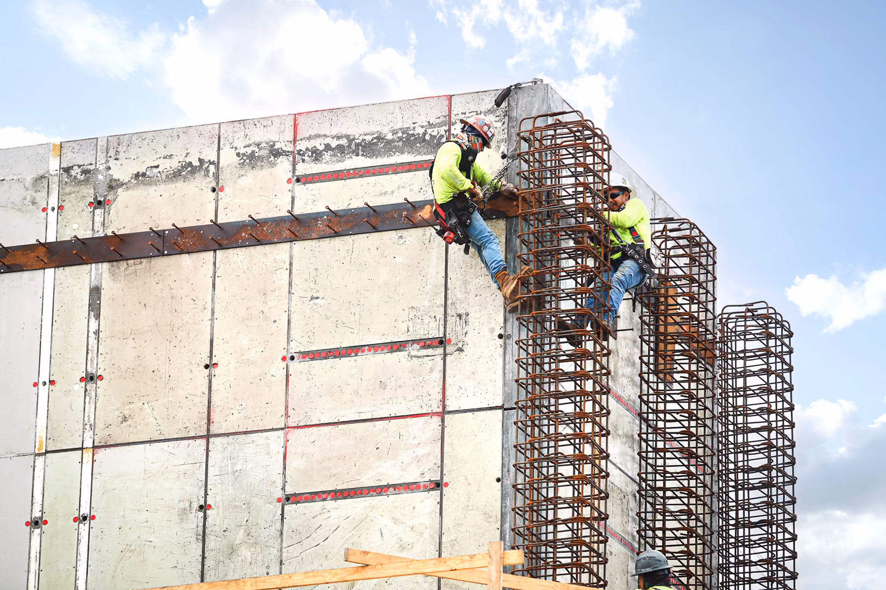 Two construction workers in safety gear working on concrete wall with metal rebar under blue sky.