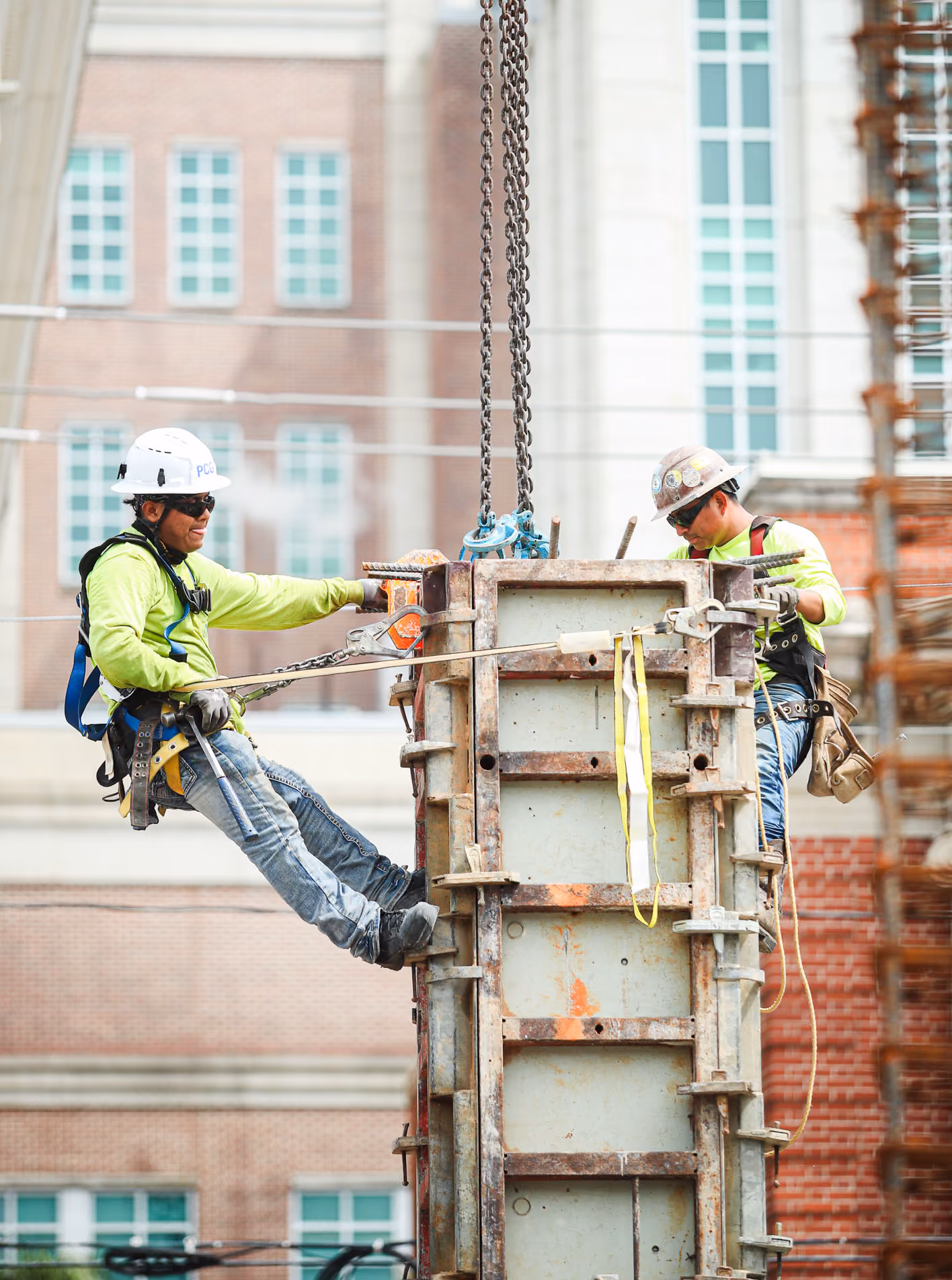 Two construction workers wearing helmets and safety harnesses working on a suspended metal structure.