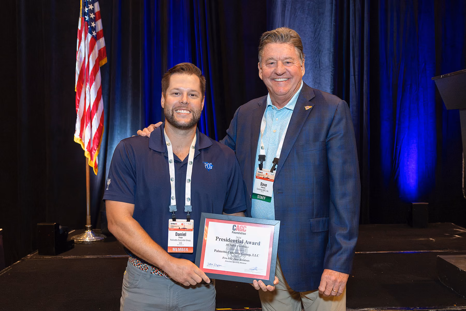 Two men smiling, one holding a framed award certificate on stage with an American flag behind.