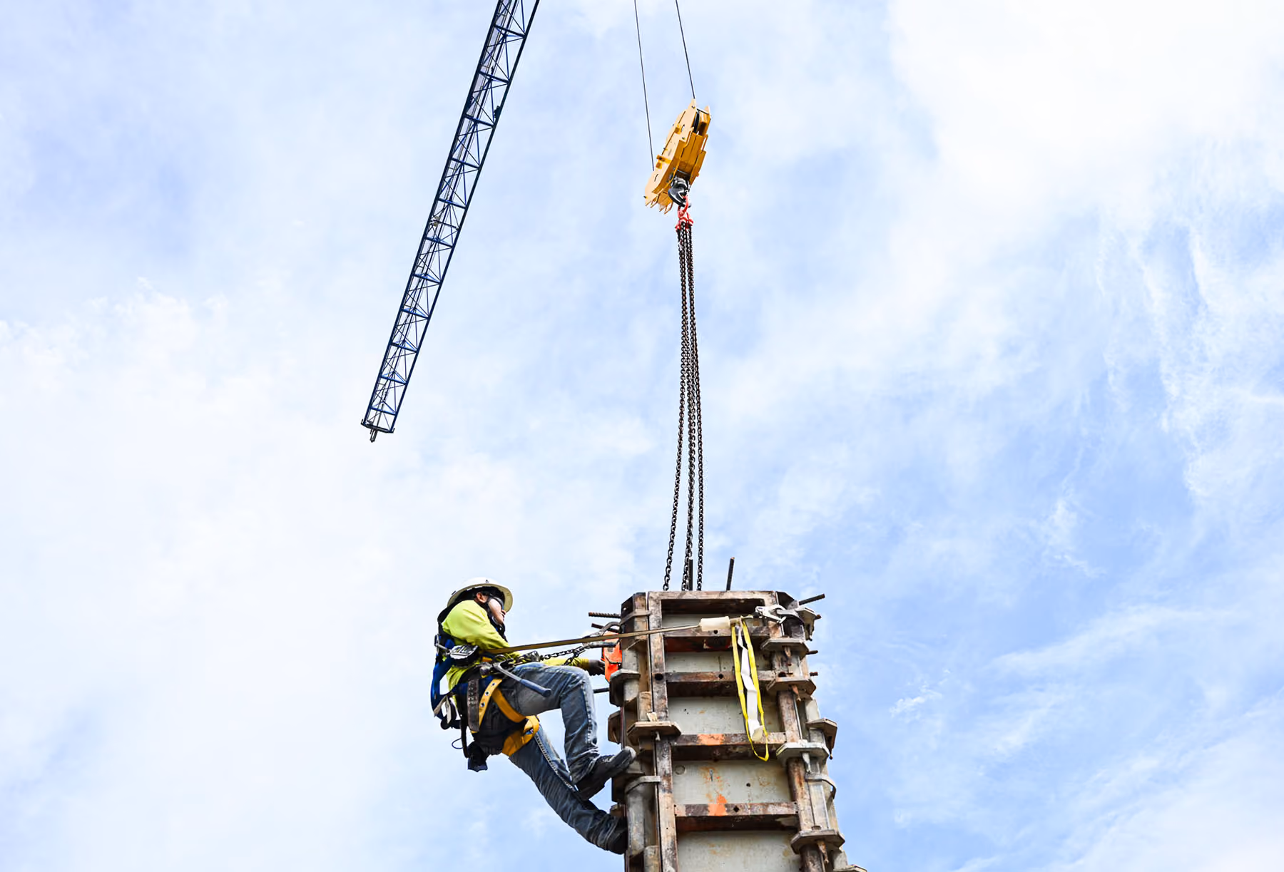 Construction worker in safety gear suspended by chains near crane against blue sky.