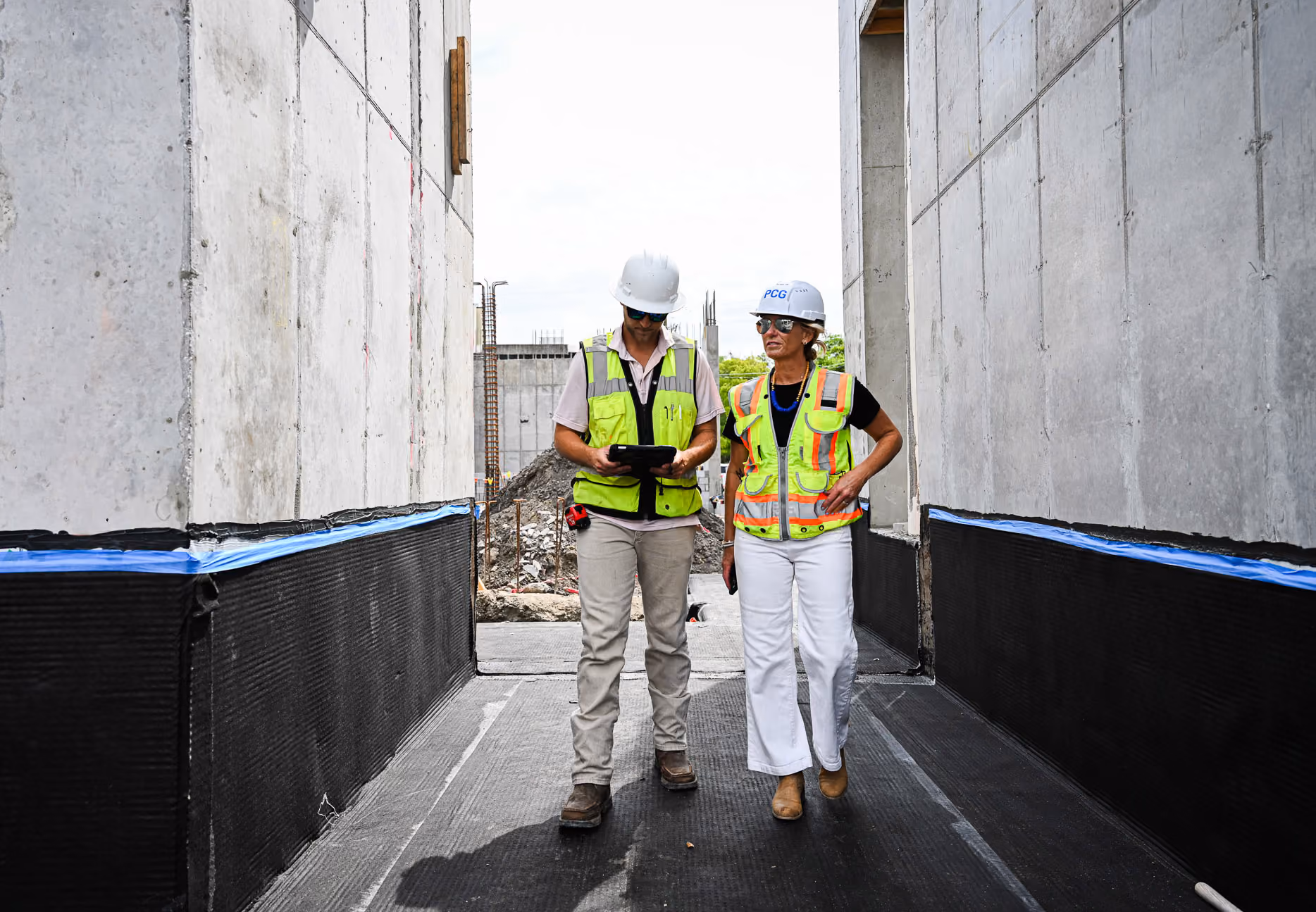Two construction workers in safety vests and helmets walking between concrete walls on a construction site.