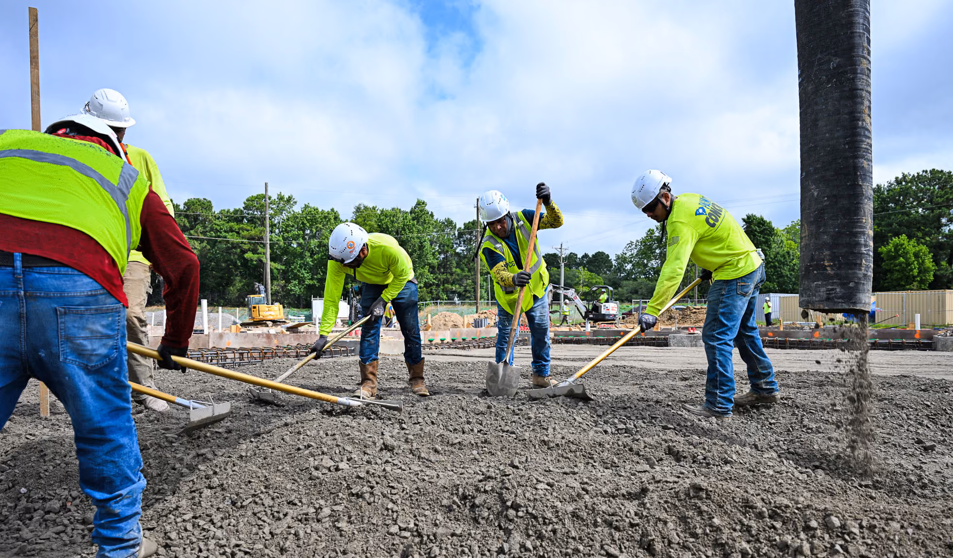 Construction workers in safety gear leveling freshly poured concrete at a job site under blue sky.