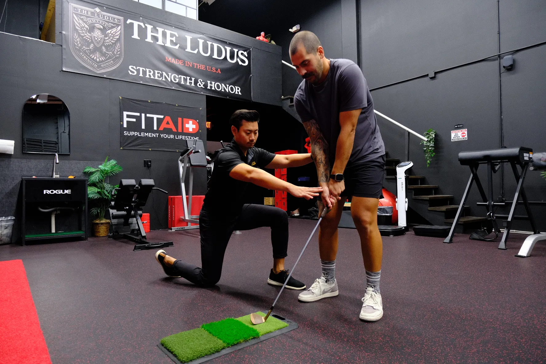 Golf instructor guiding a man’s golf swing indoors on a practice mat with gym equipment around.