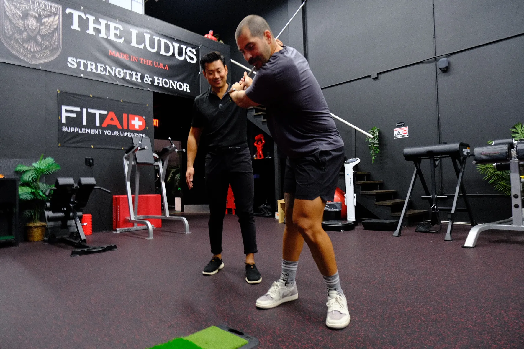 Coach instructing a man with a golf club in an indoor gym with exercise equipment.