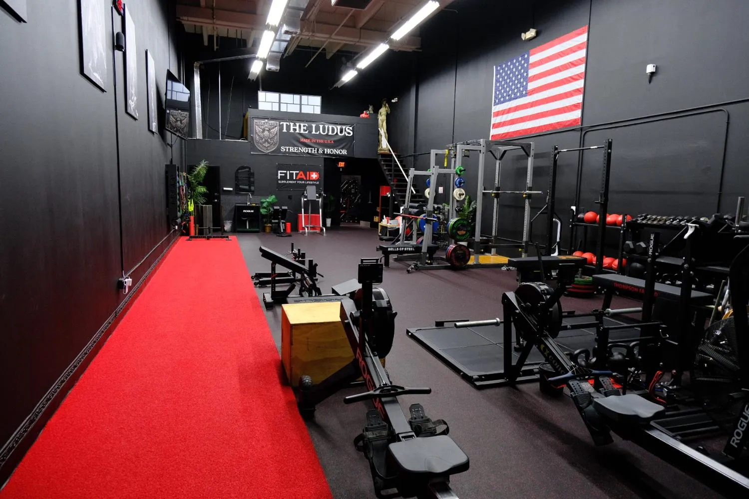 Interior of a gym with rowing machines, weight racks, a red running track, and an American flag on the wall.