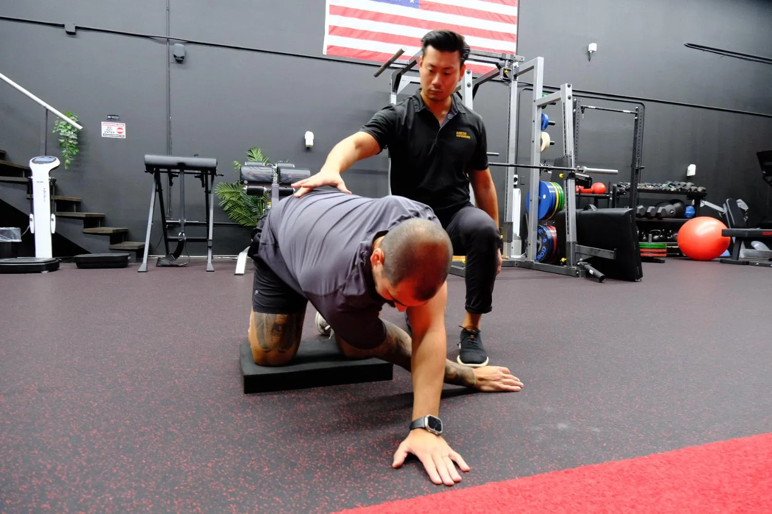 Trainer assists man stretching while kneeling on a mat in a gym with exercise equipment.
