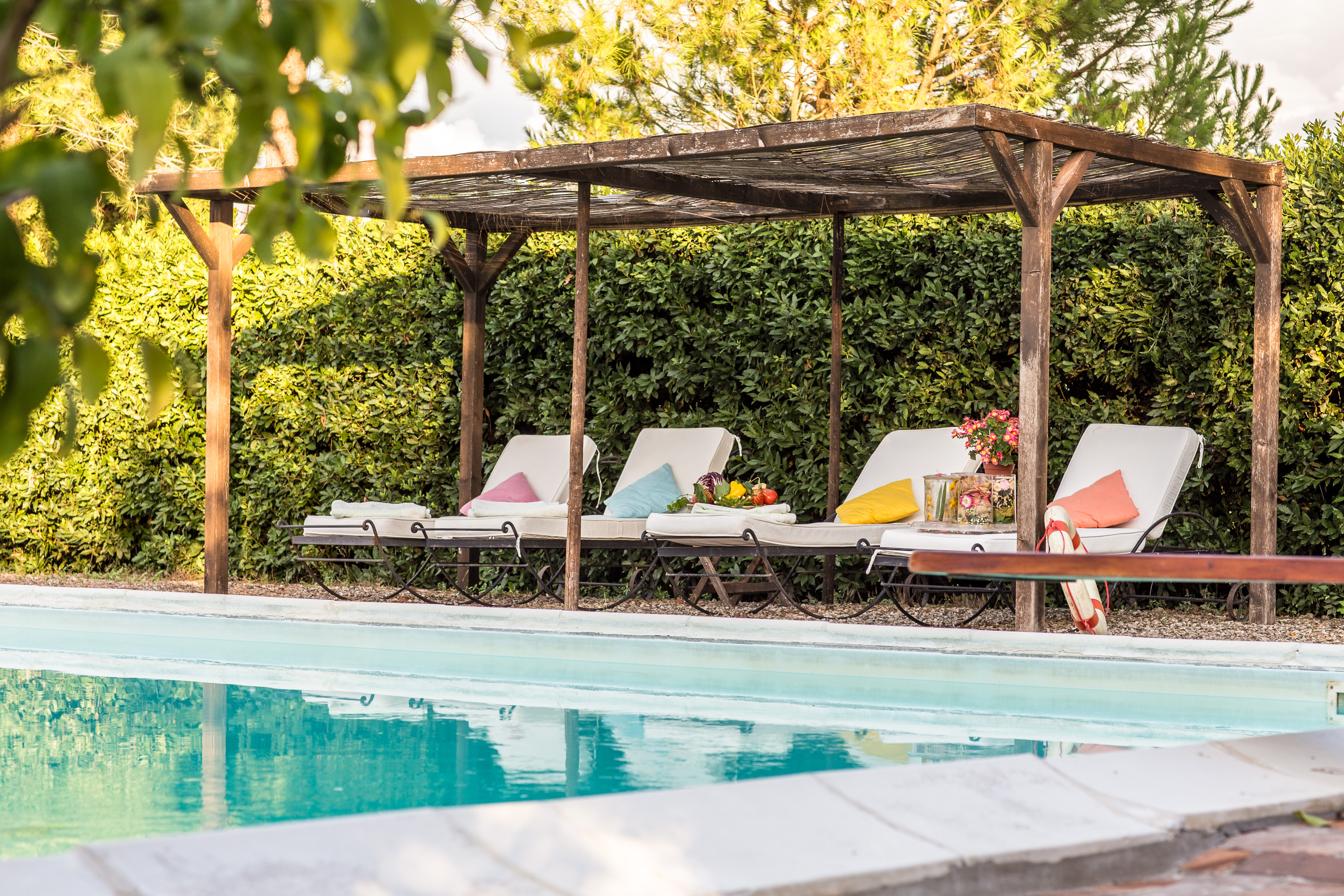Four cushioned lounge chairs with colorful pillows under a wooden pergola beside a swimming pool, Castello Higo, Tuscany.