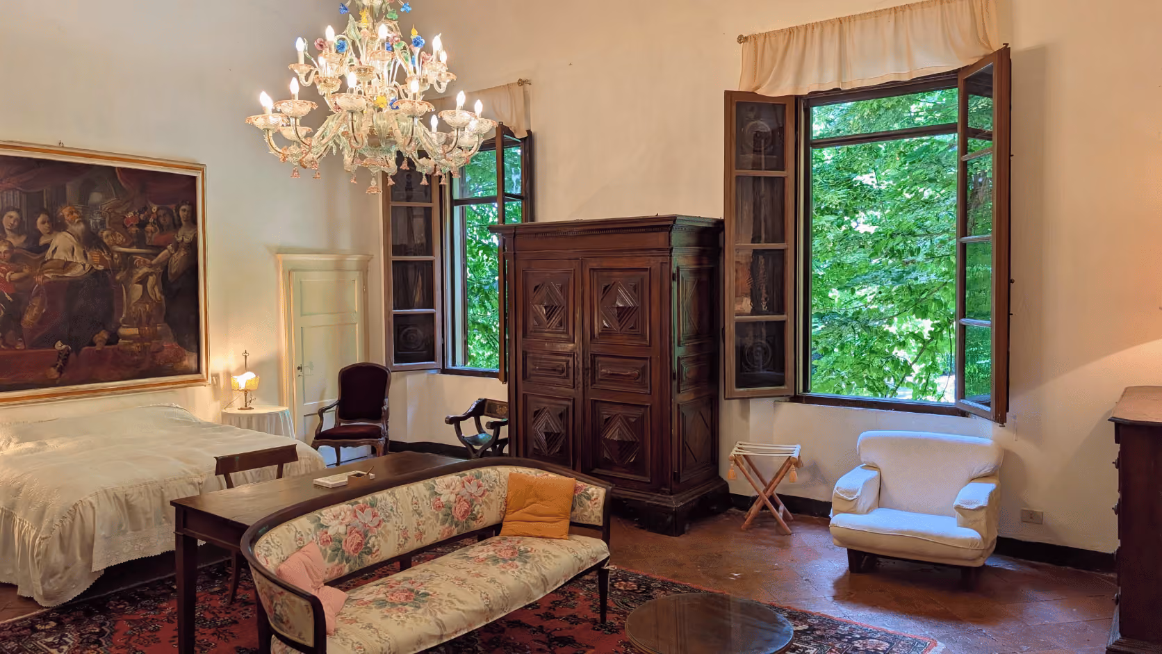 Una Camera with floral sofa, wooden armoire, chandelier, and open windows showing green trees in Castello Higo rooms