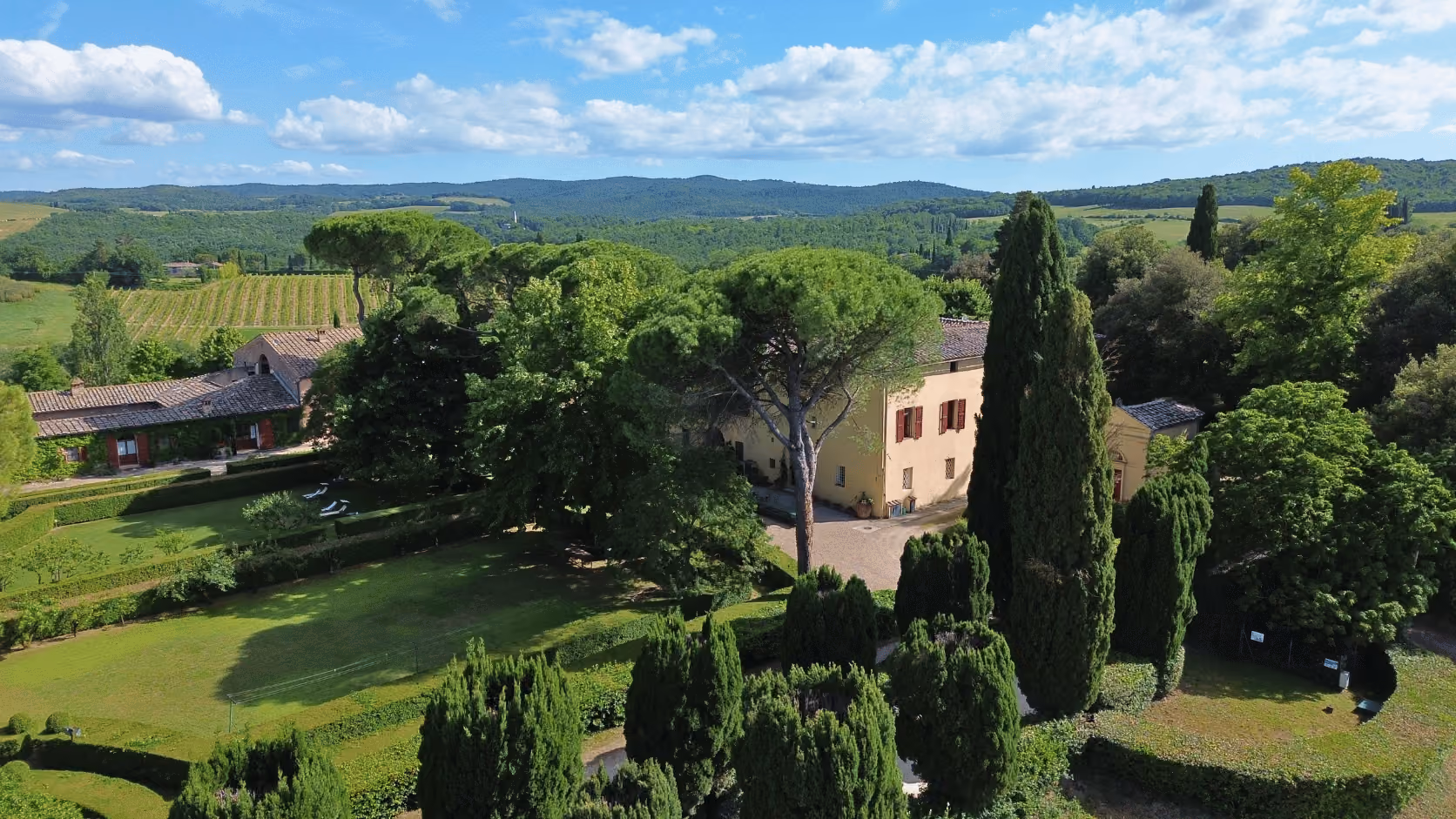 Countryside estate at Castello Higo, Tuscany, with vineyard, tall trees, hedges, and a tan building under a partly cloudy sky.