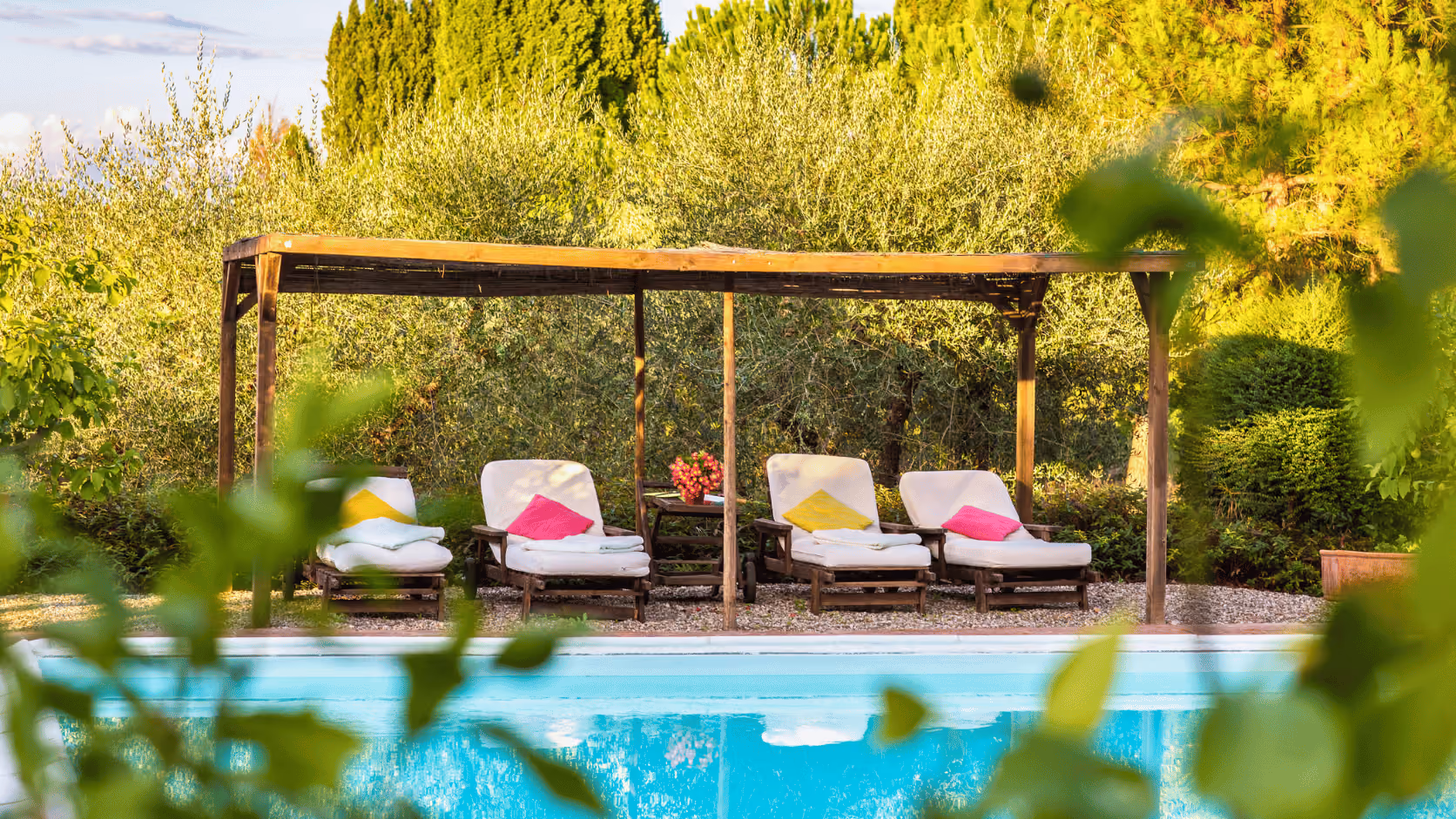 Poolside lounge at Castello Higo with cushioned chairs beneath a wooden pergola, surrounded by lush greenery