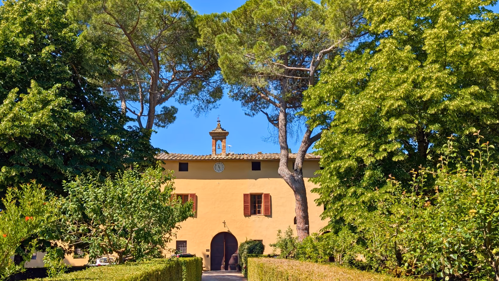 Yellow covered fasade of Castello Higo with a clock and bell tower surrounded by green trees and bushes under a blue sky.