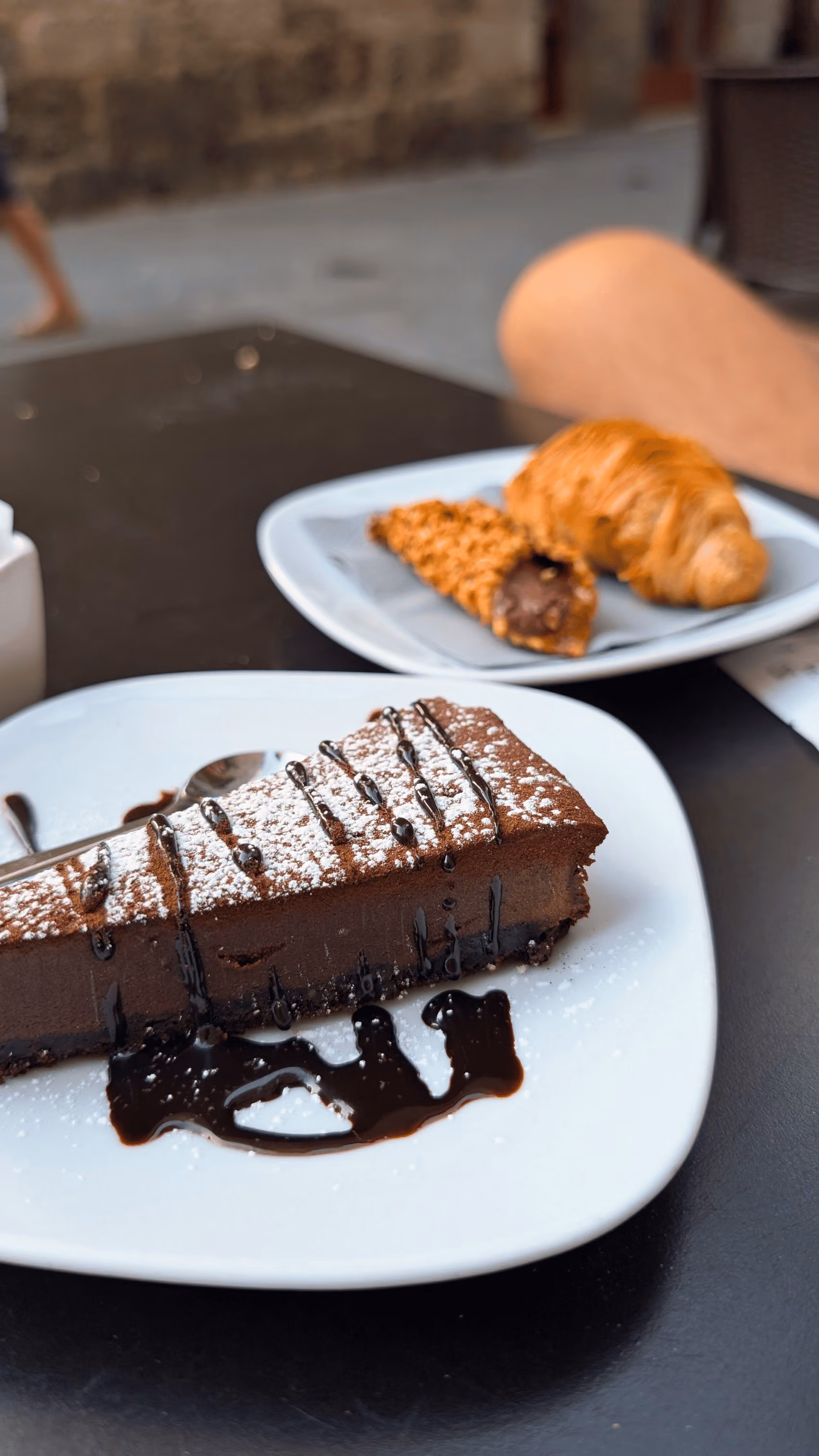 Slice of chocolate cake with powdered sugar and chocolate drizzle on a white plate with pastry in the background, Castello Higo, Tuscany.