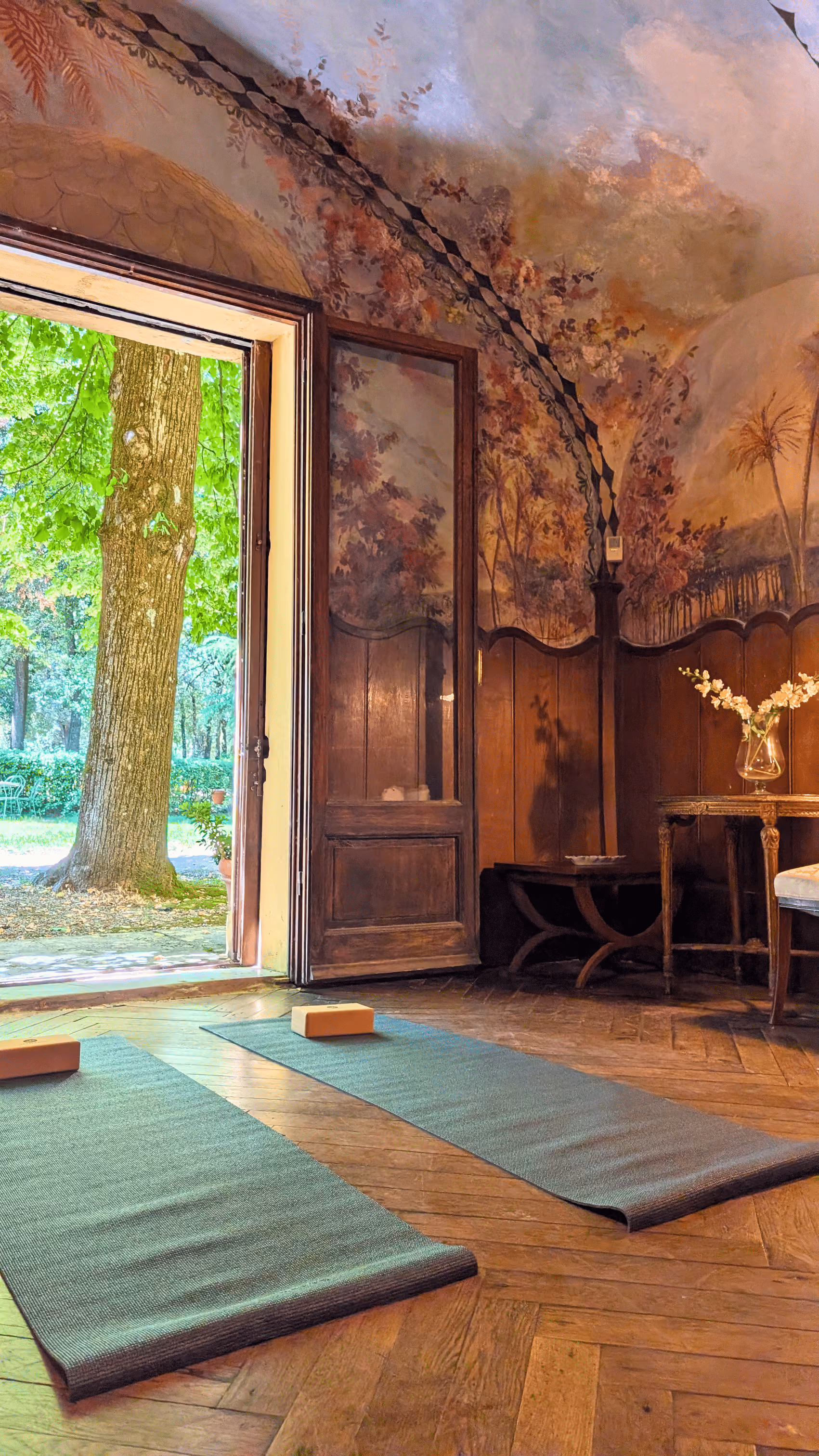 Two yoga mats with blocks on a wooden floor inside the fresco-covered yoga studio at Castello Higo, Tuscany, with an open door to the garden.