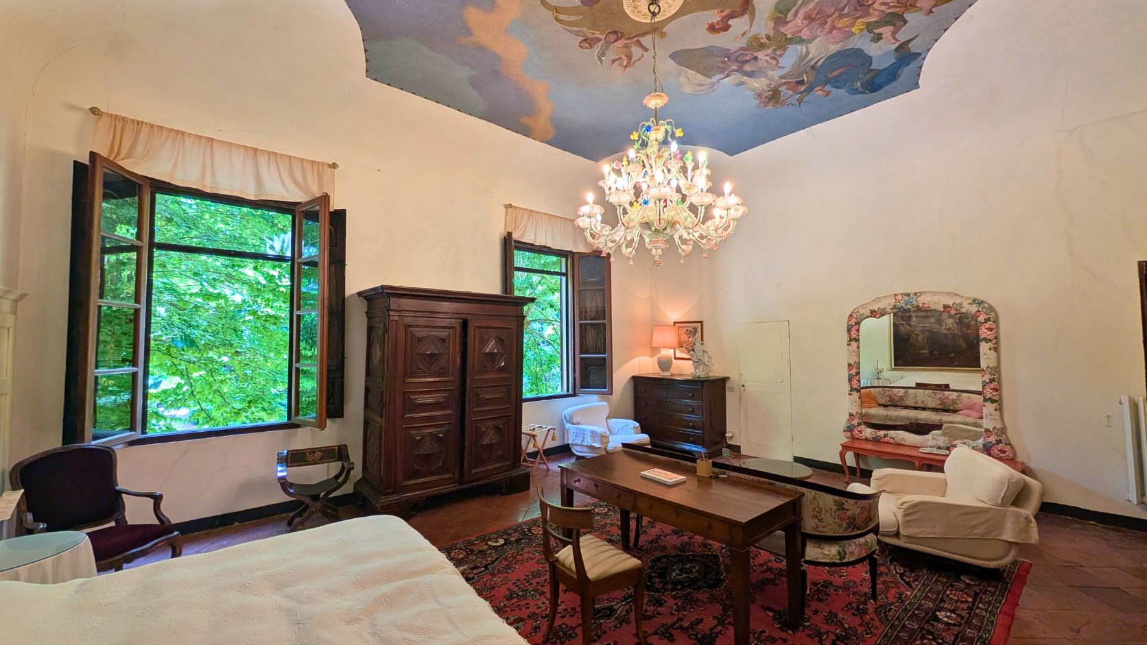 Spacious vintage room with open windows, wooden furniture, an ornate chandelier, painted ceiling, and large mirror, Castello Higo, Tuscany.