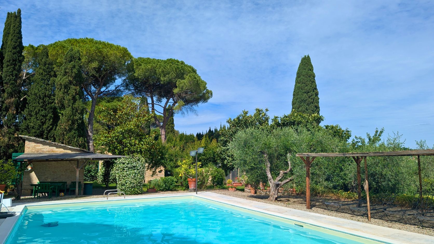 Clear blue swimming pool with green trees, shrubs, and a shaded seating area in the background, Castello Higo, Tuscany.