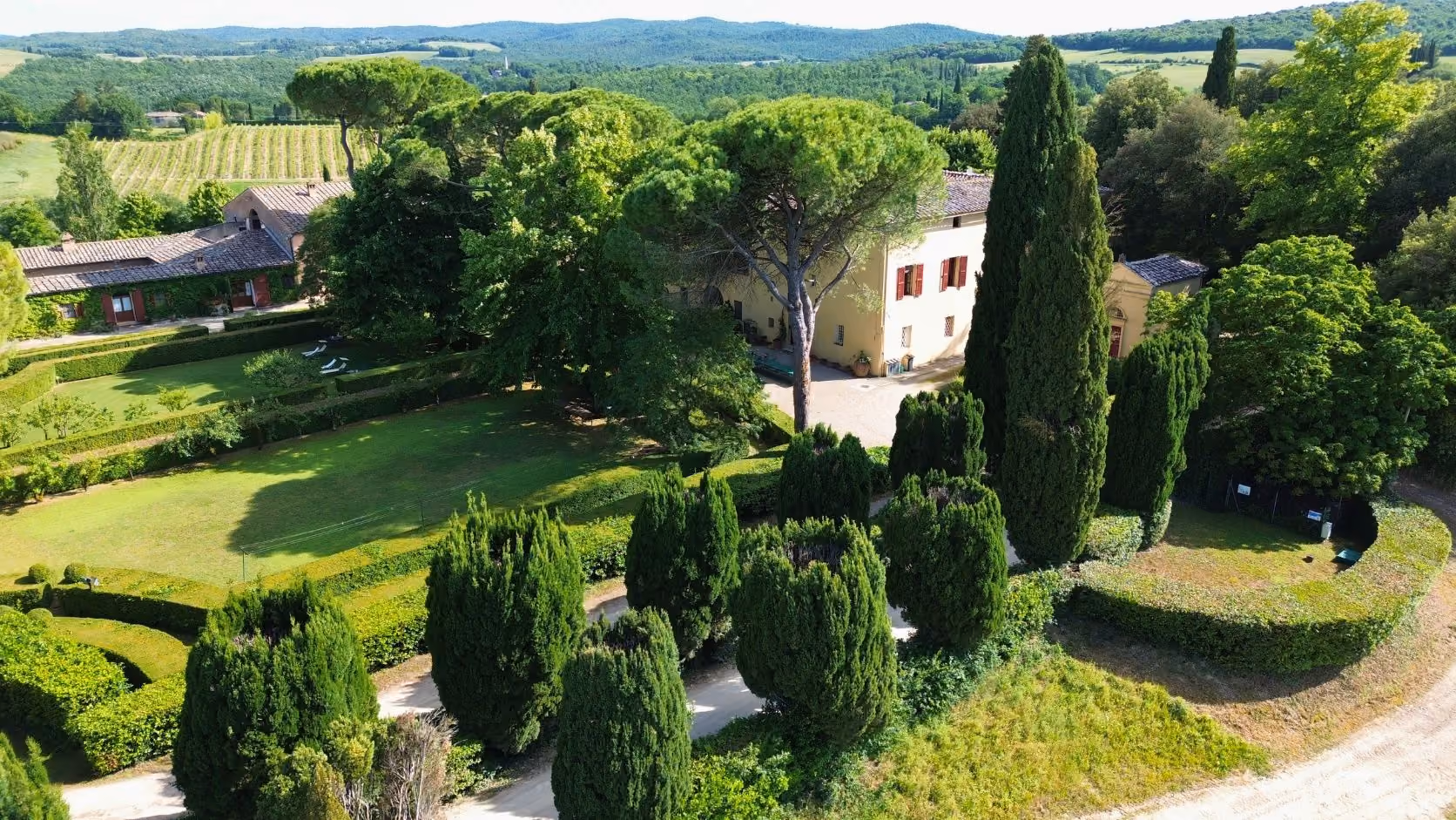 Aerial view of a yellow villa surrounded by tall trees, manicured gardens, and rolling green hills, Castello Higo, Tuscany.