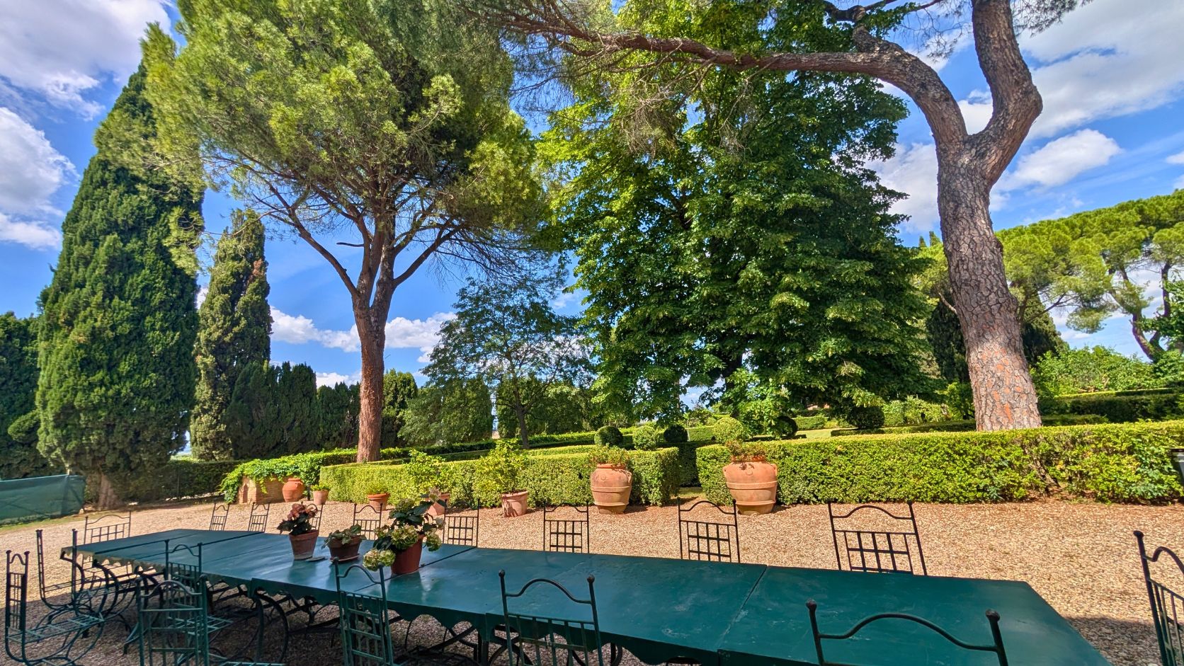 Long outdoor green metal table and chairs set on gravel under large trees in a sunny garden, Castello Higo, Tuscany.