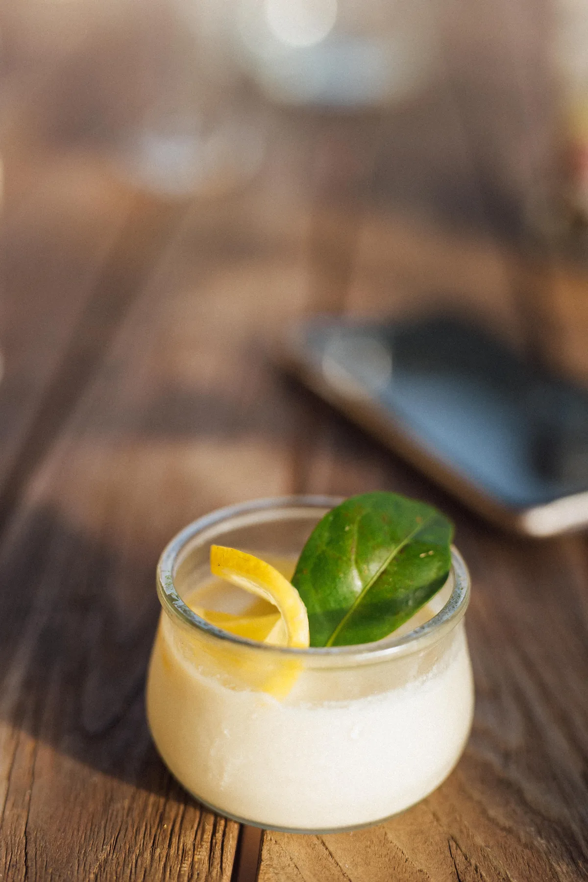 A glass jar with a creamy drink garnished with a lemon peel and green leaf on a wooden table, Castello Higo, Tuscany.