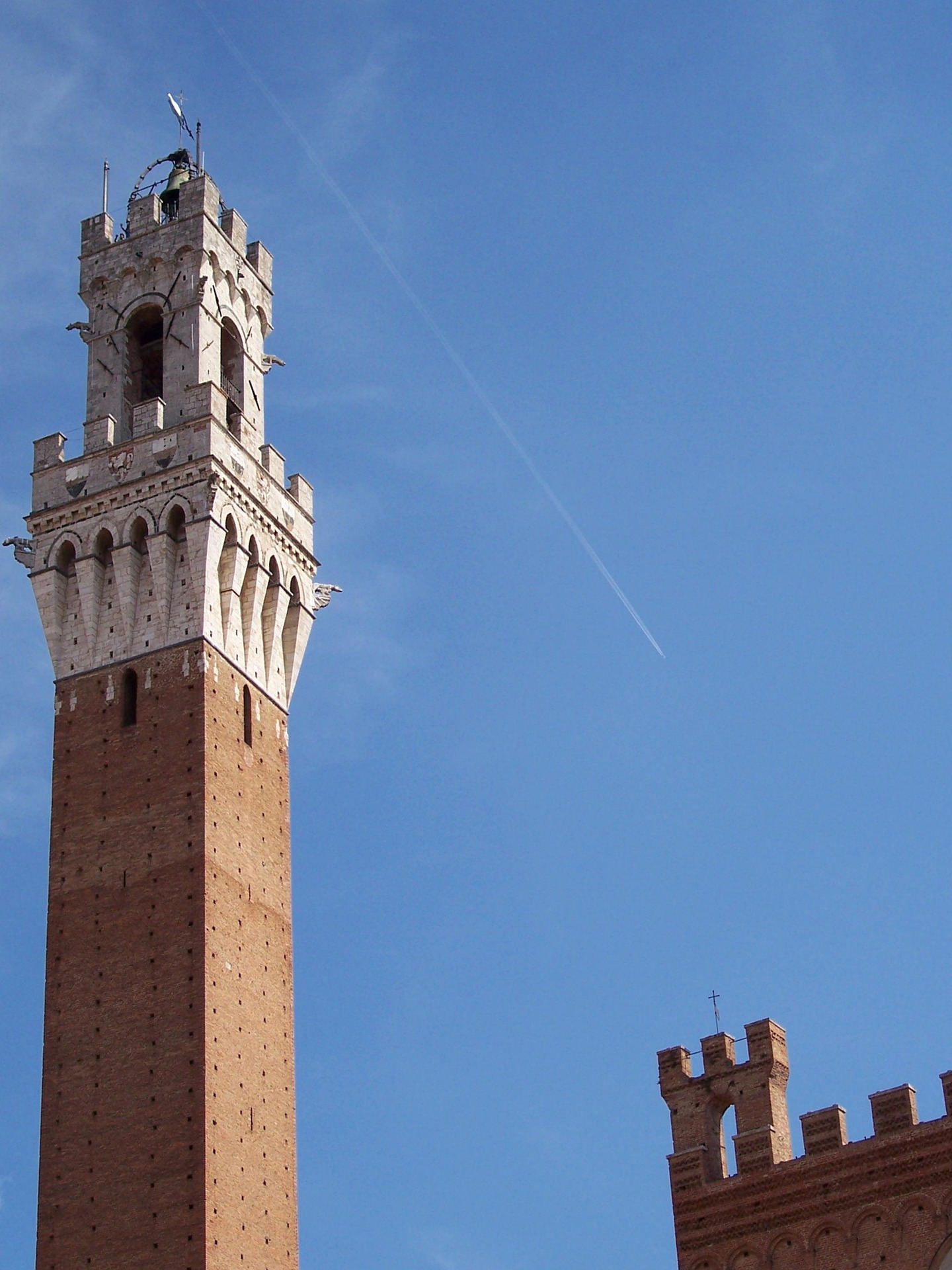 Siena’s medieval Torre del Mangia, a tall brick tower crowned with ornate stonework, rises against a clear blue sky beside a faint airplane contrail.