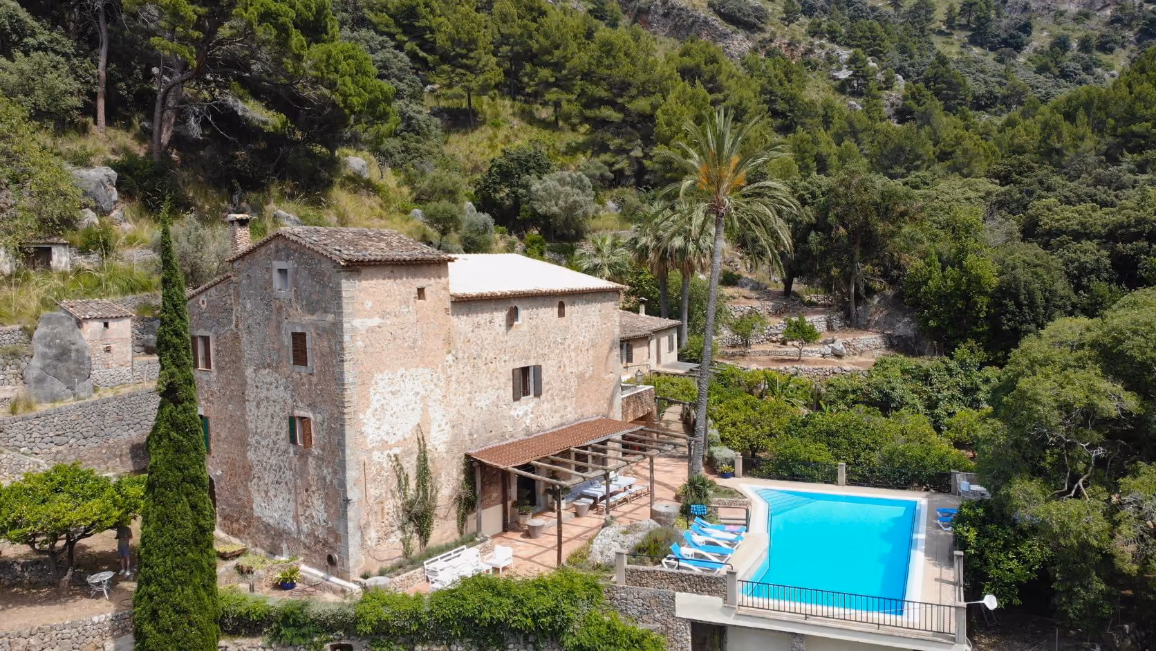 Stone house with tiled roof beside a blue swimming pool surrounded by lush green trees and hills, Finca Higo, Mallorca.