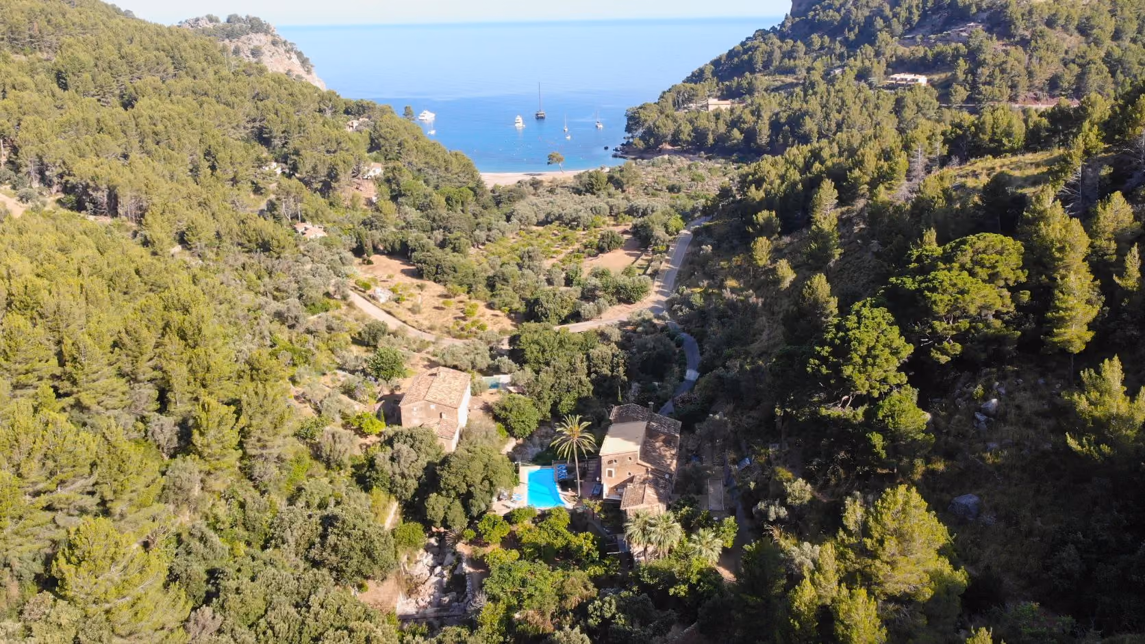 Aerial view of houses with a pool surrounded by trees near a cove with boats on calm blue water, Finca Higo, Mallorca.
