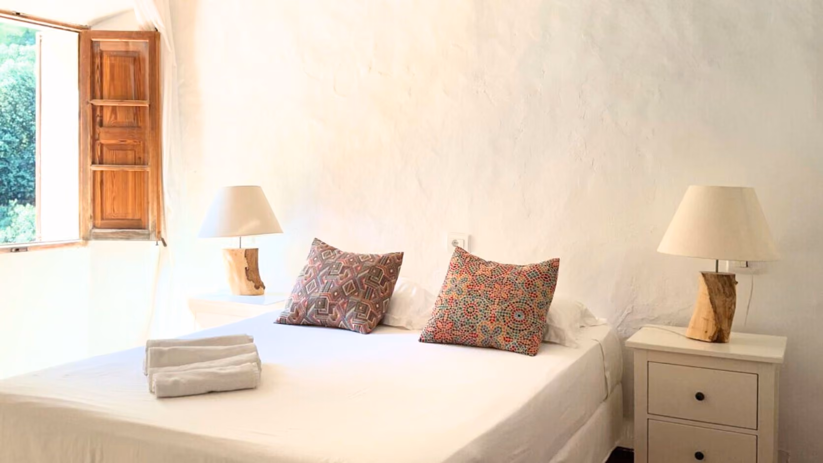 Minimalist bedroom with white bed, patterned pillows, wooden lamps, and an open window showing greenery, Finca Higo, Mallorca.
