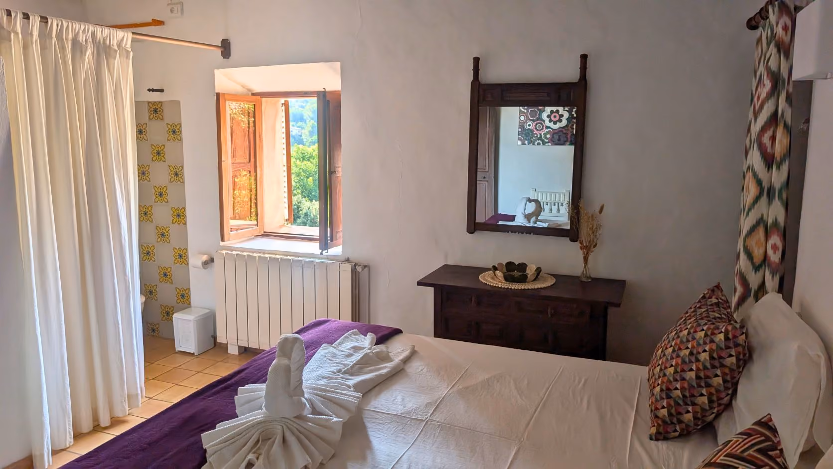 Cozy bedroom with white bed, folded towel swan, colorful pillows, window with view, and wooden dresser with mirror, Finca Higo, Mallorca.