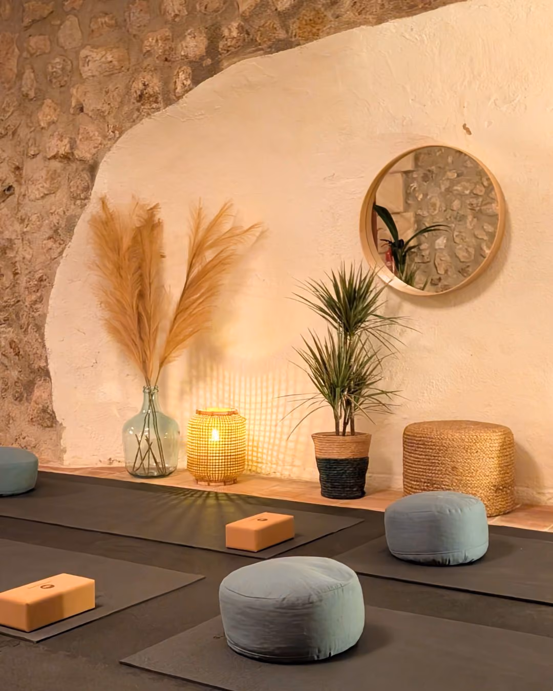 Yoga mats with cushions and blocks in the yoga shala of Finca Higo, Mallorca, with stone wall, plants, lamp, and round mirror.