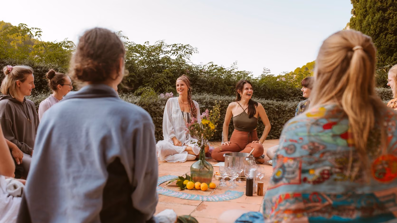 Finca Higo yoga participants in Mallorca sitting in a circle outdoors, smiling and chatting around a vase with flowers and fruit.