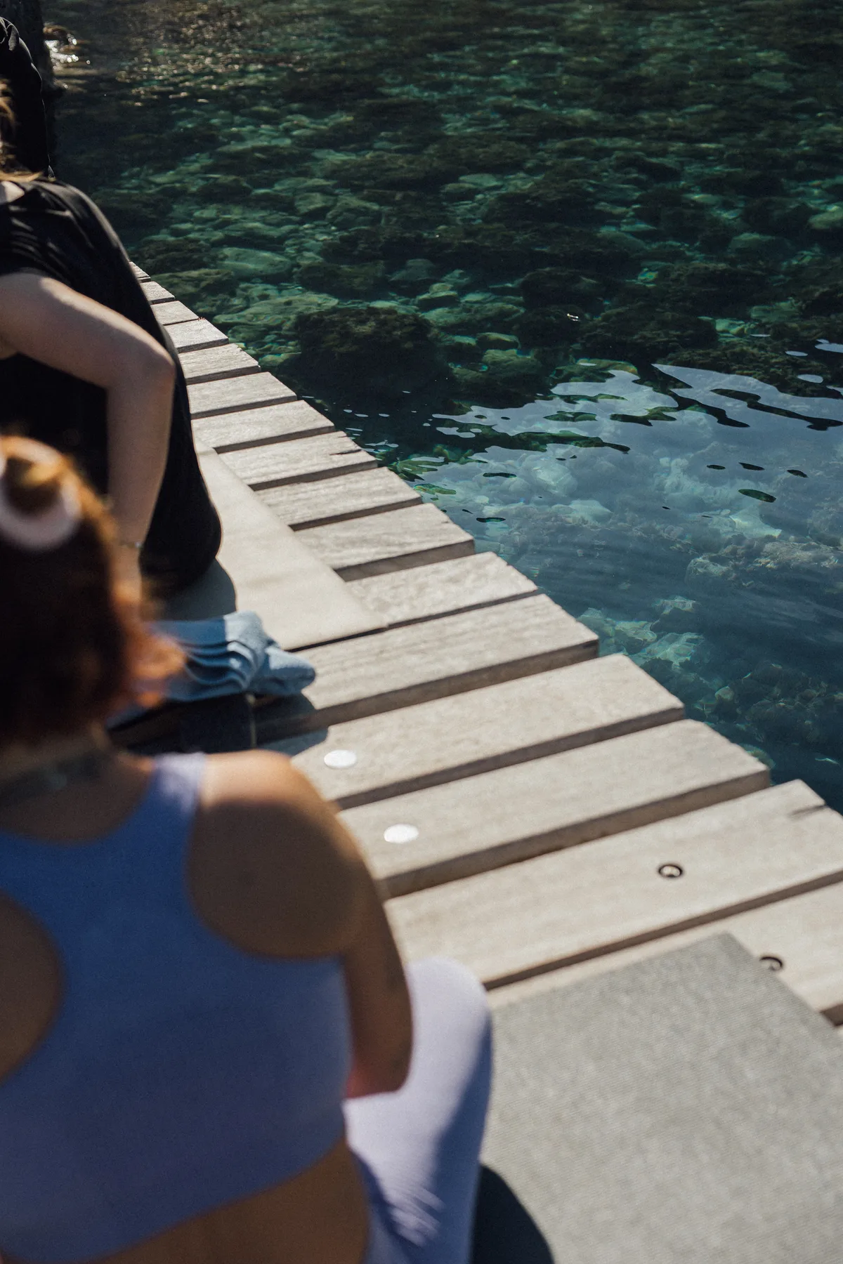 Two people sitting on a wooden dock beside clear water with rocks visible below, Finca Higo, Mallorca.