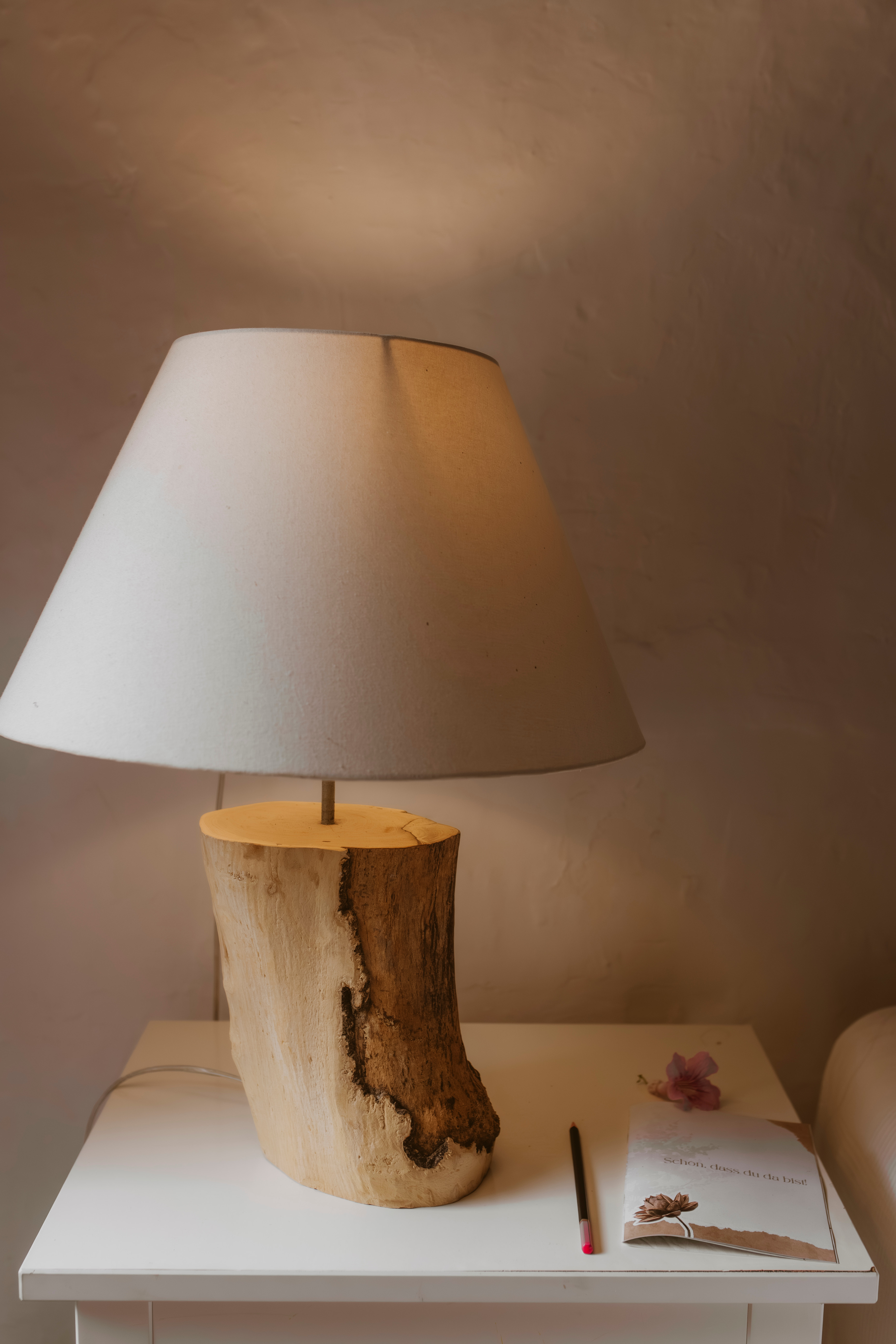 Table lamp with a wooden base and beige shade on a white nightstand with a pencil and notebook, Finca Higo, Mallorca.