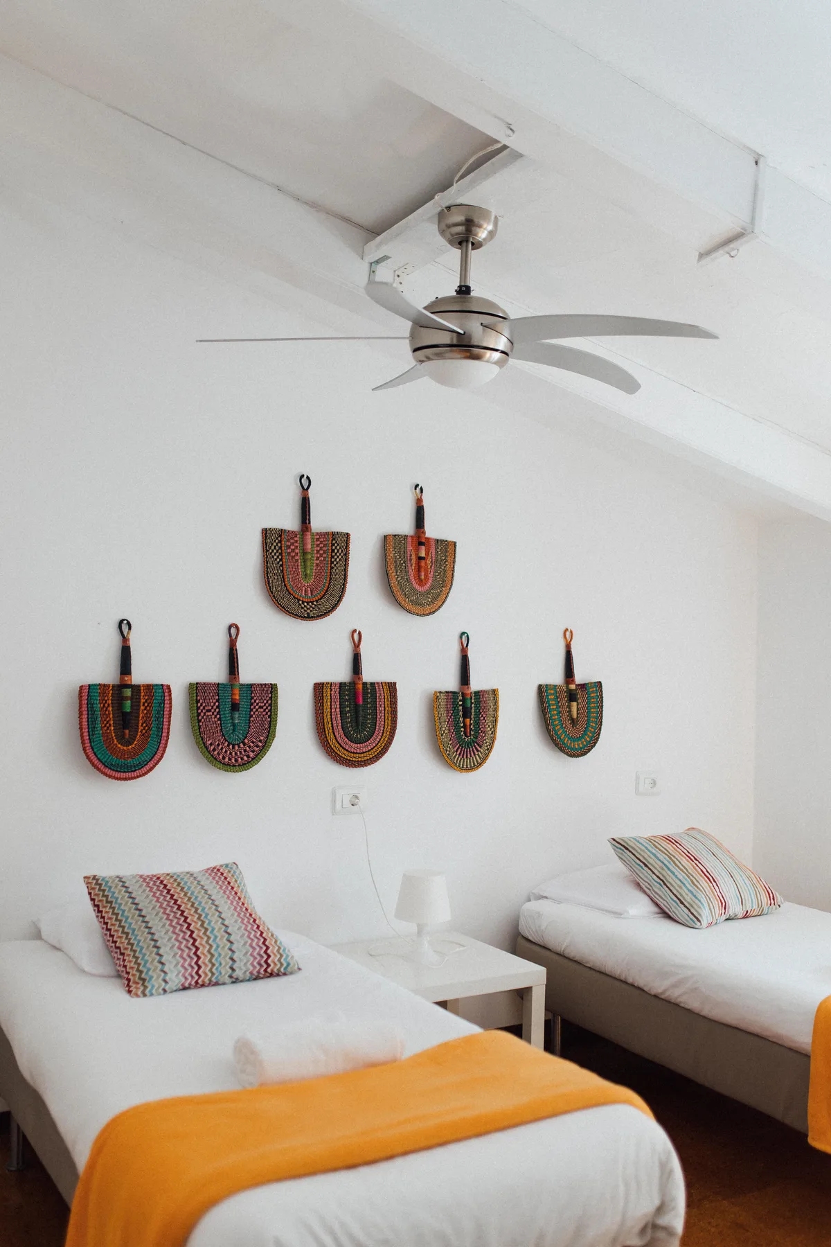 Minimalist bedroom with two single beds, colorful pillows, orange blankets, and woven wall decor, Finca Higo, Mallorca.