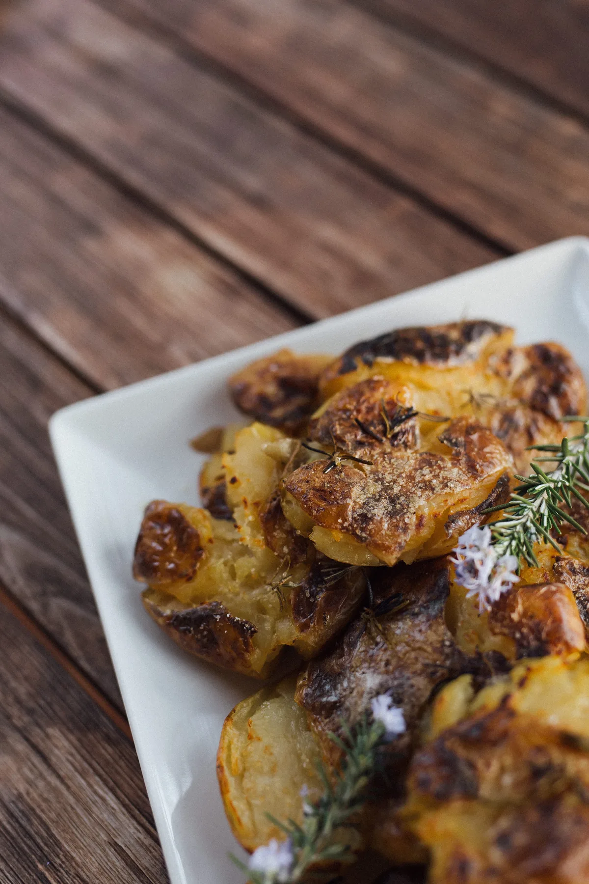 Crispy roasted smashed potatoes garnished with rosemary on a white plate over a wooden surface, Castello Higo, Tuscany.