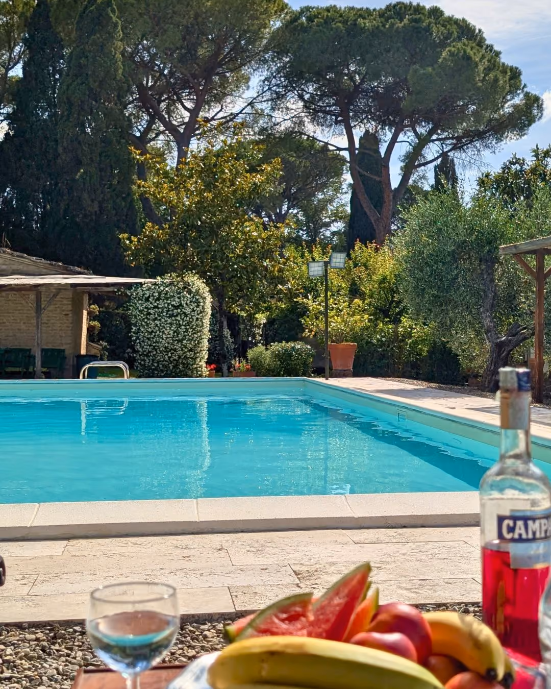 Outdoor swimming pool with clear water surrounded by trees and a table holding fruit and a bottle, Castello Higo, Tuscany.