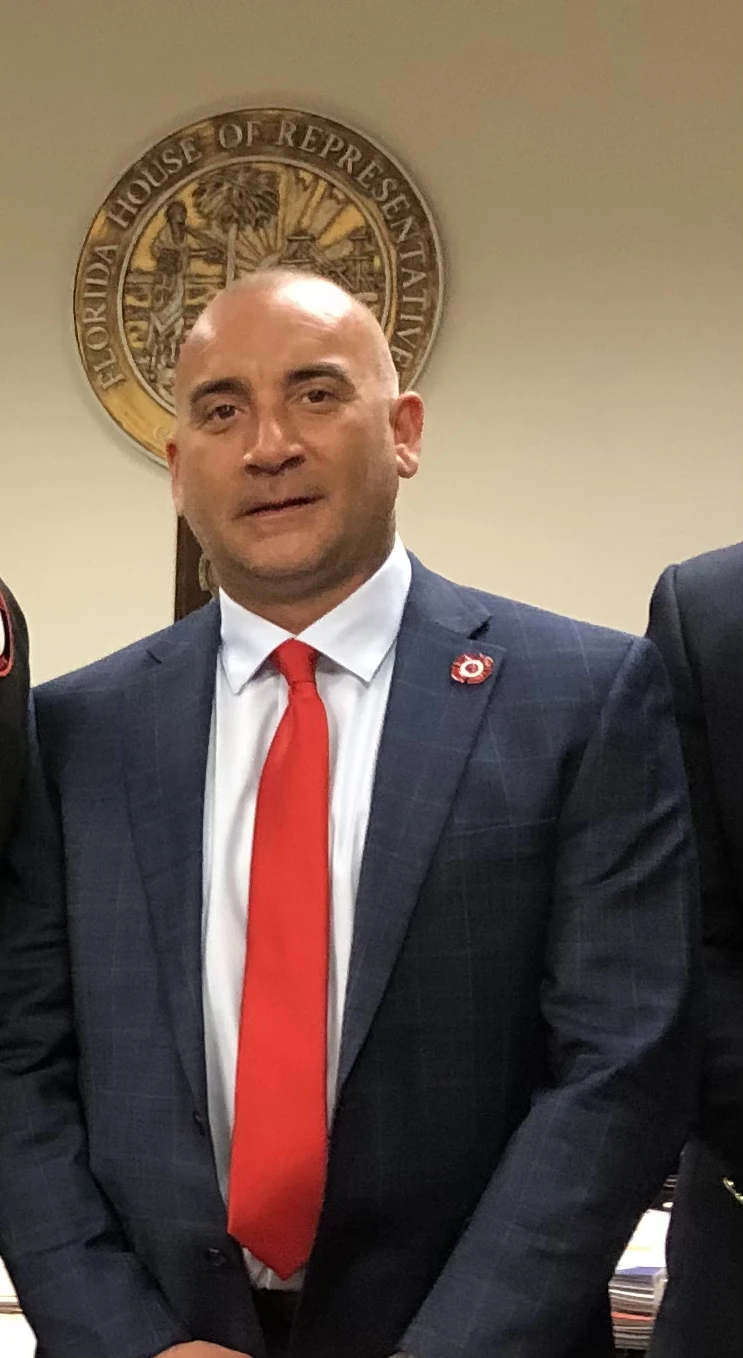 Man in dark suit, red tie, and white shirt standing in front of Florida House of Representatives seal.