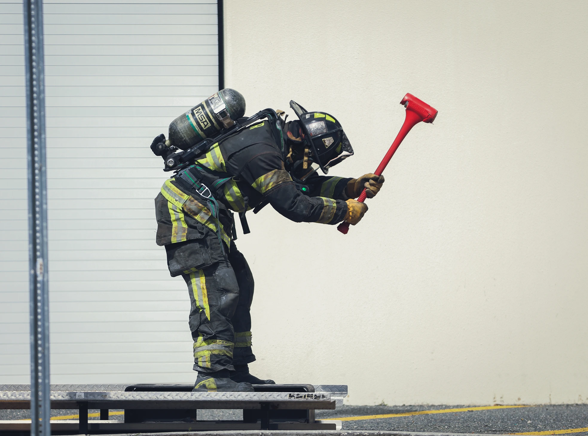 Firefighter in full gear raising a red axe, standing on a raised platform outdoors.