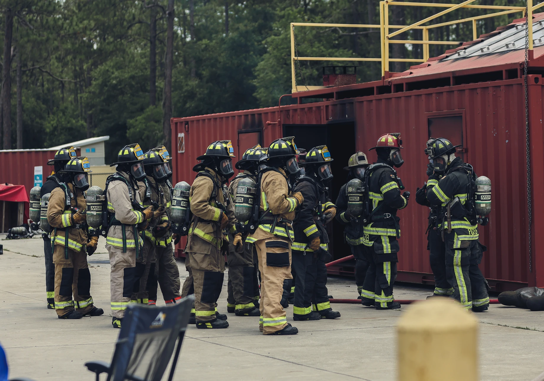 Group of firefighters in full gear lined up outside a red training container near forest.