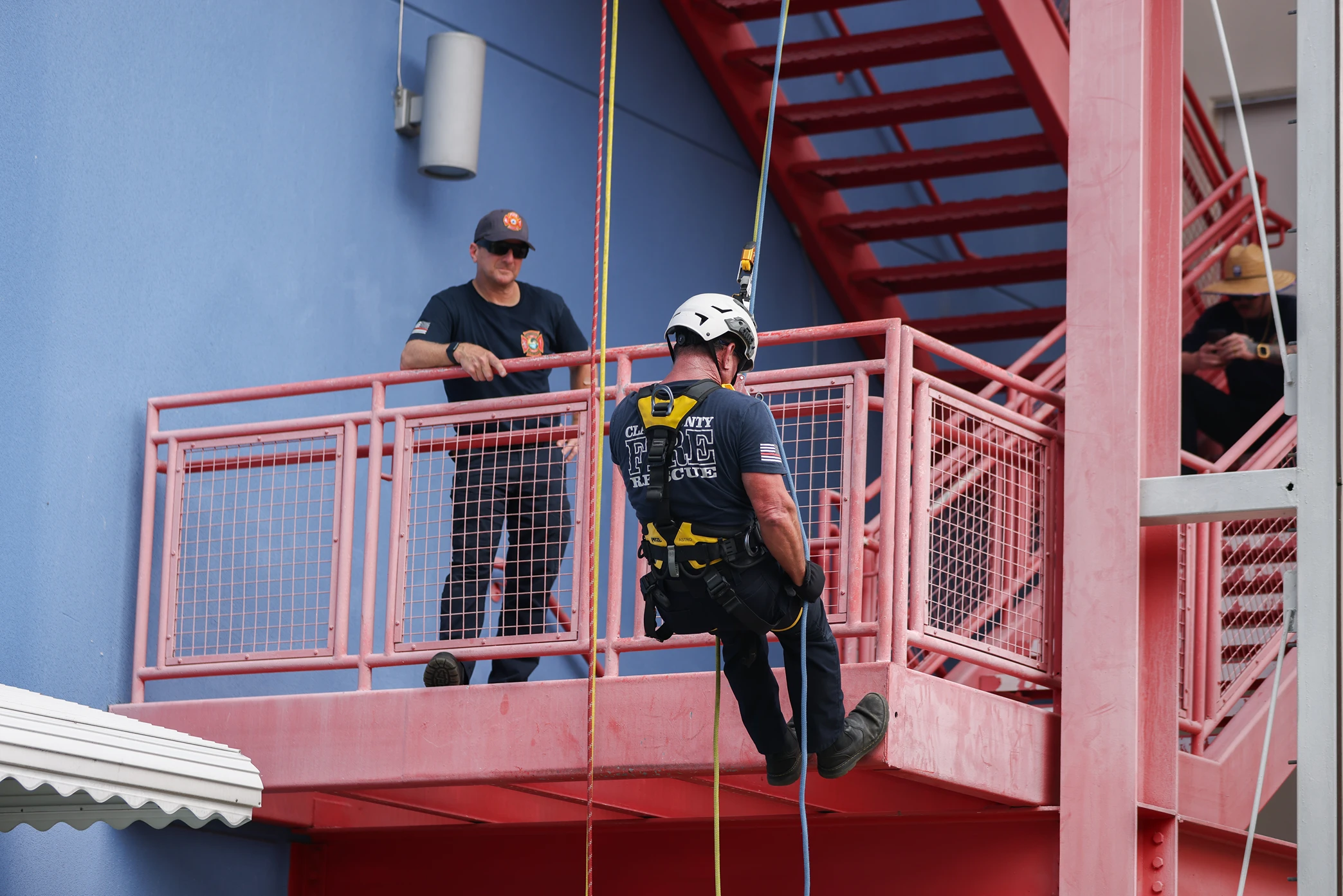 Firefighter in helmet and harness rappels down red staircase platform while another watches nearby.