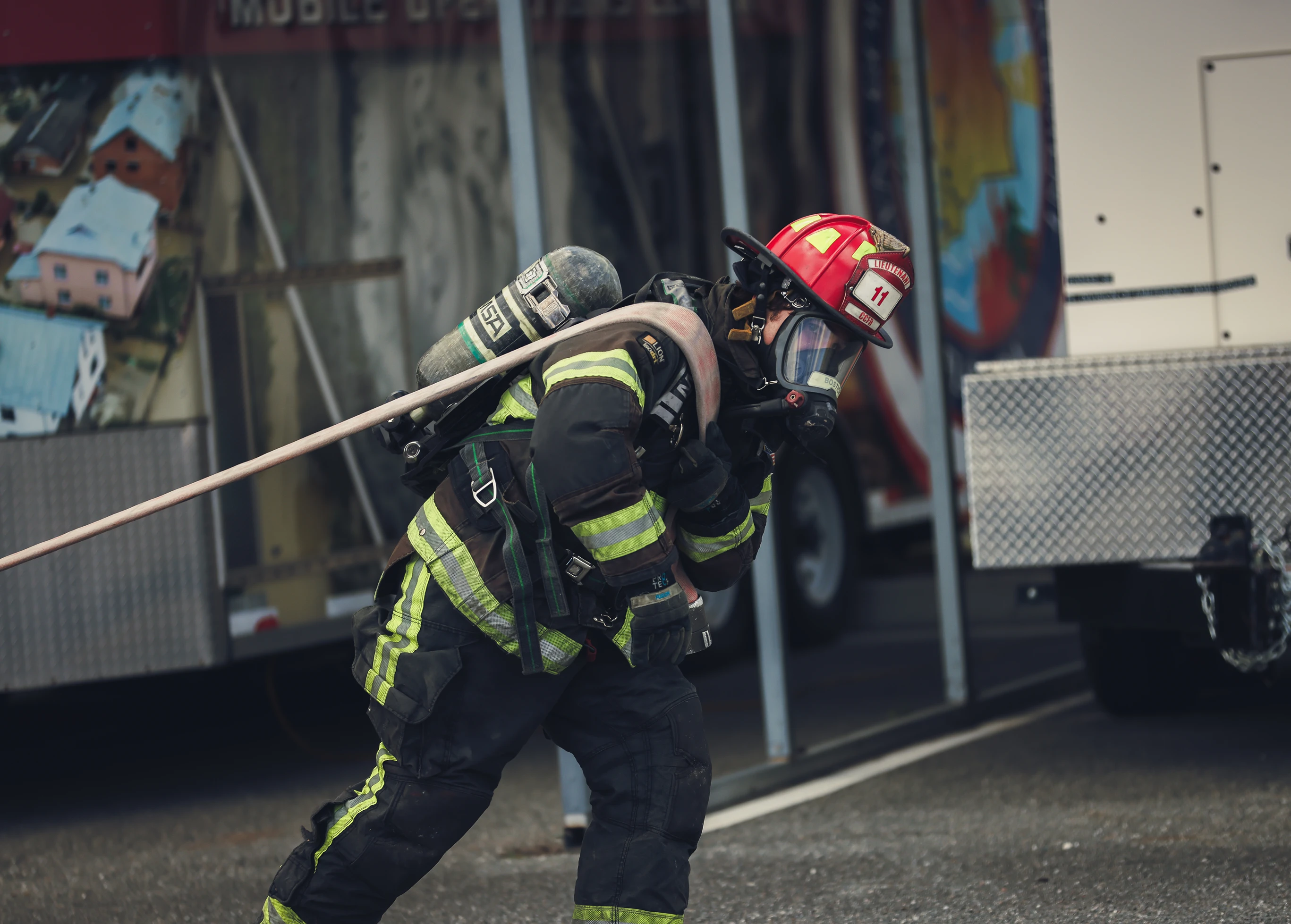 Firefighter in full gear pulling a fire hose near emergency vehicles.