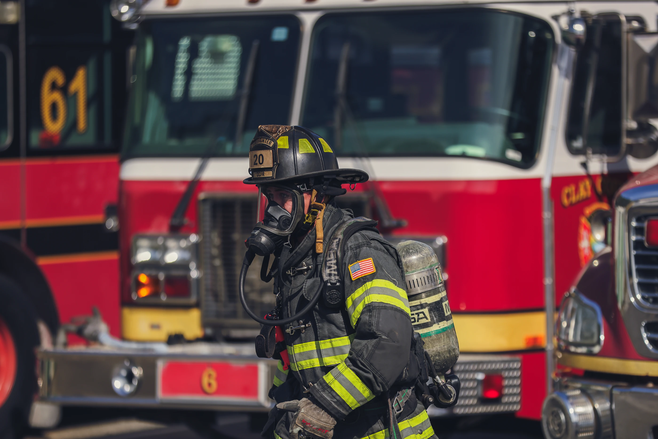Firefighter in full gear with oxygen tank standing in front of red fire trucks.