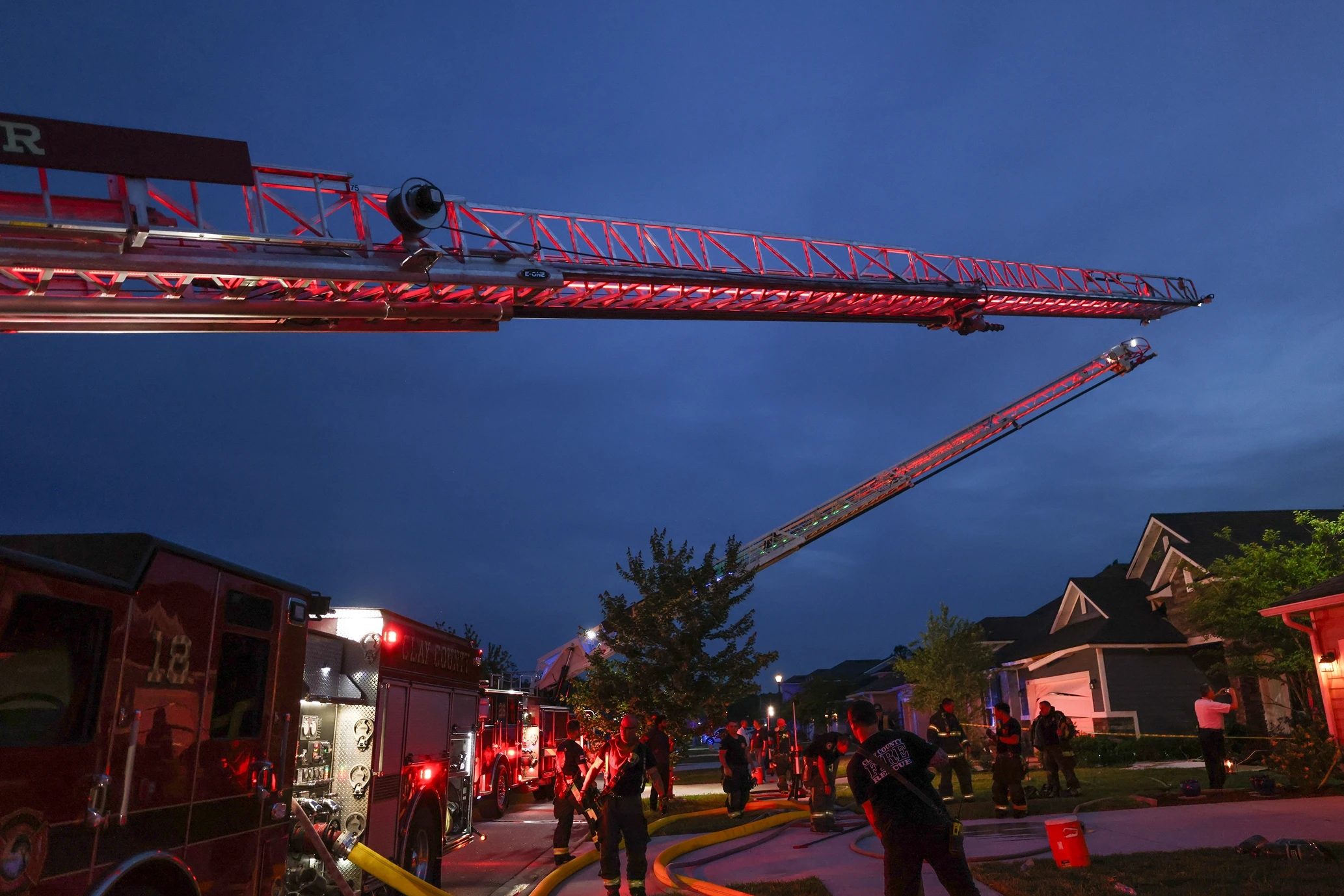 Firefighters and trucks with extended ladders responding to an emergency at a residential neighborhood at dusk.