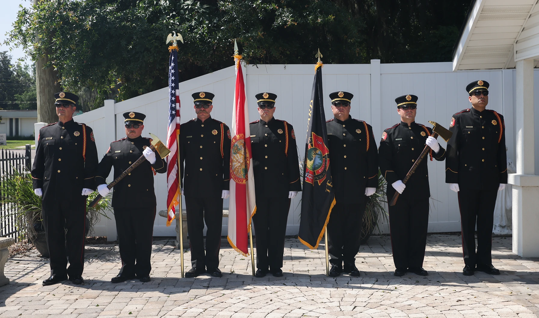 Seven firefighters in formal uniforms stand in a line holding flags and axes for a ceremony.
