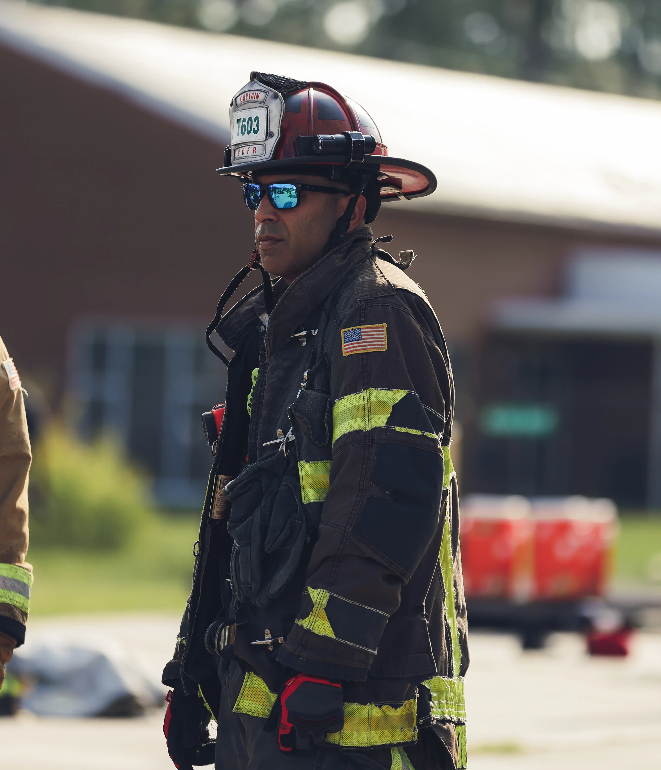 Firefighter wearing helmet labeled captain and reflective gear, standing outdoors with gloves and sunglasses.