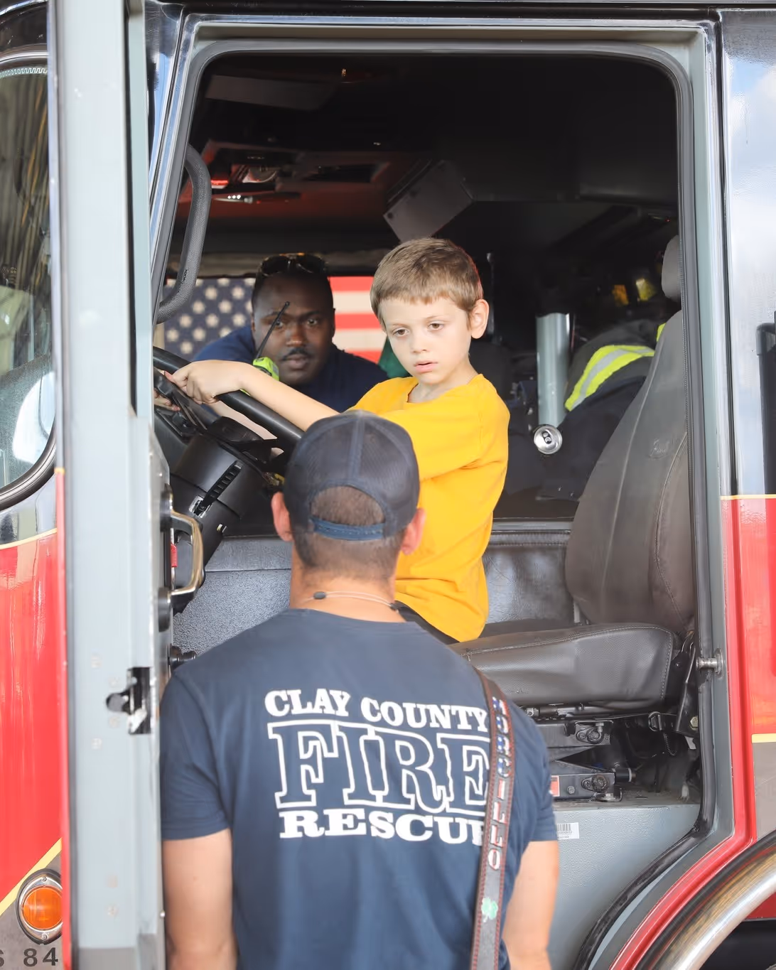 Boy in a yellow shirt sits in fire truck driver's seat with firefighter talking to him.