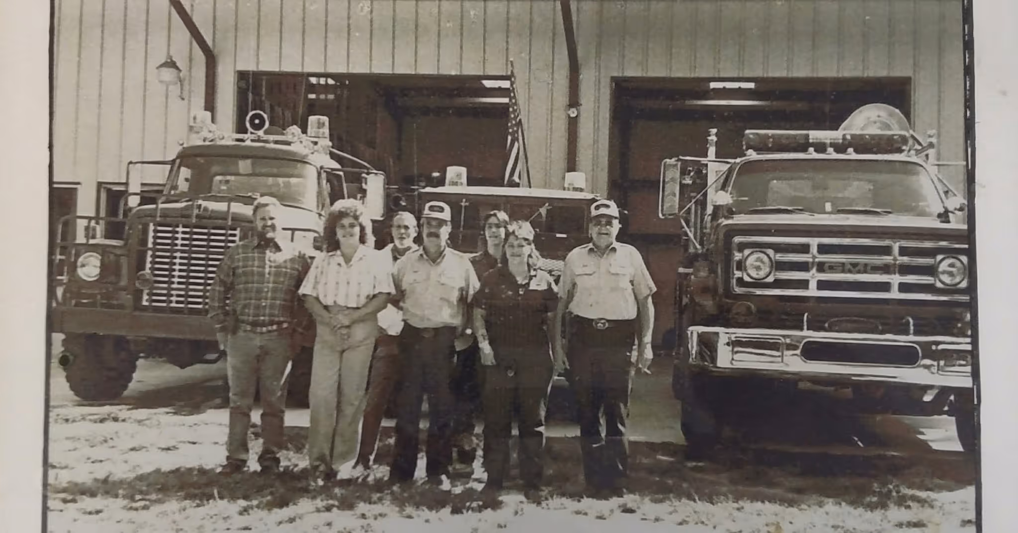Seven fire department personnel standing in front of two fire trucks inside a fire station.
